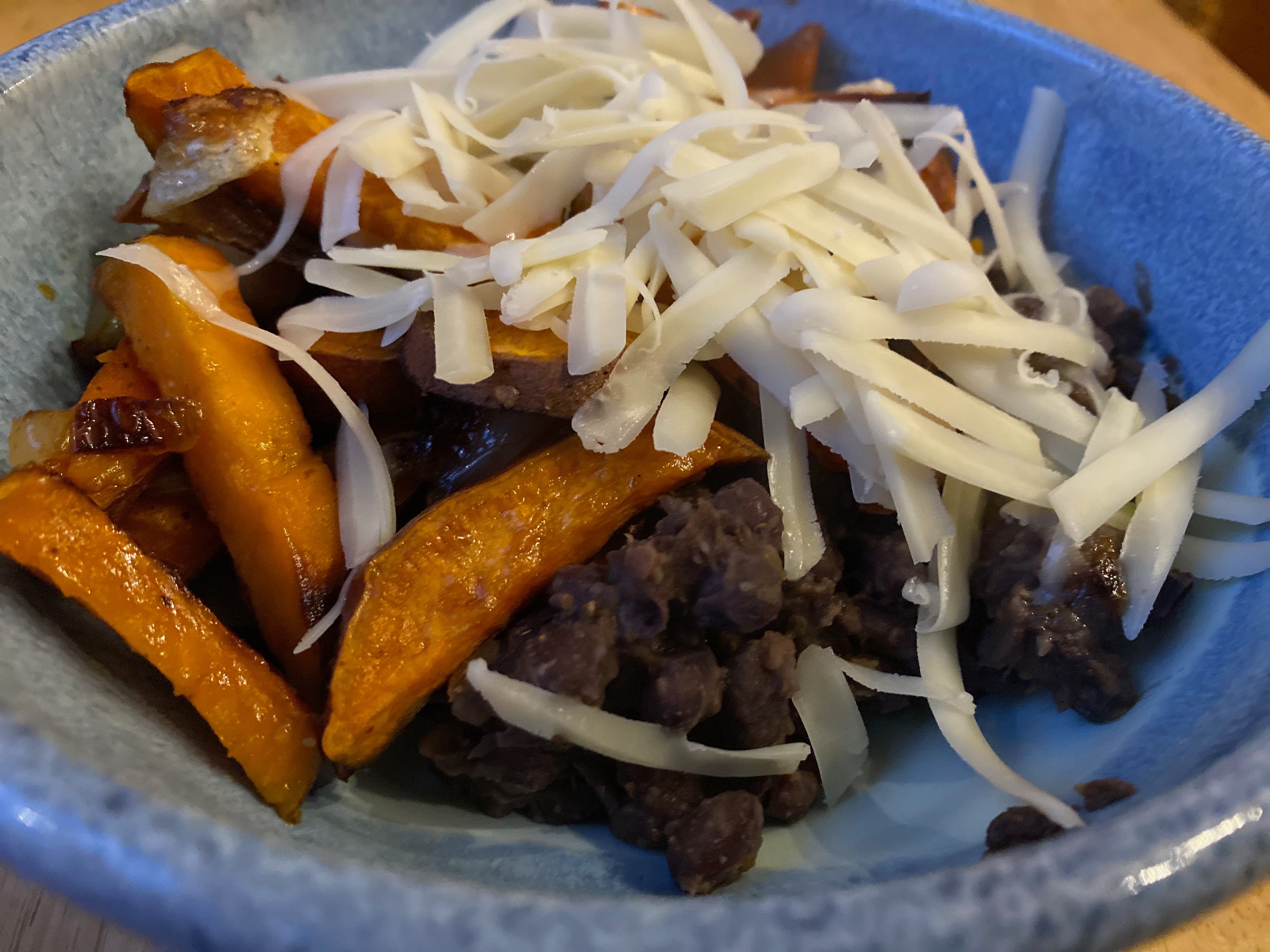 Closeup of a blue ceramic bowl filled with refried black beans, several roasted sweet potato wedges, and a pile of grated cheese.