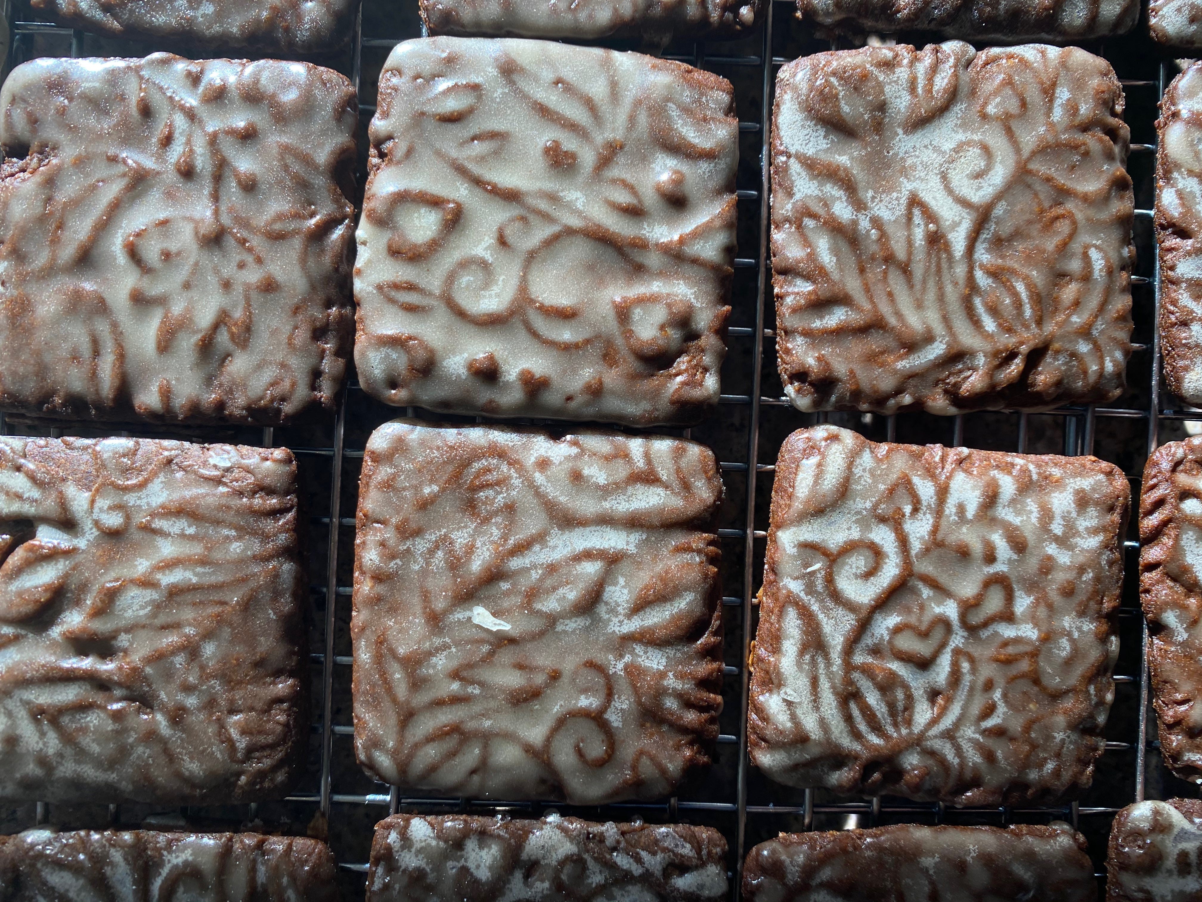A closeup of six square gingerbread cookies. Each cookie is imprinted with a pattern of leaves, flowers, and vines. They’re glazed with a semi-transparent white glaze, so the pattern is visible underneath.