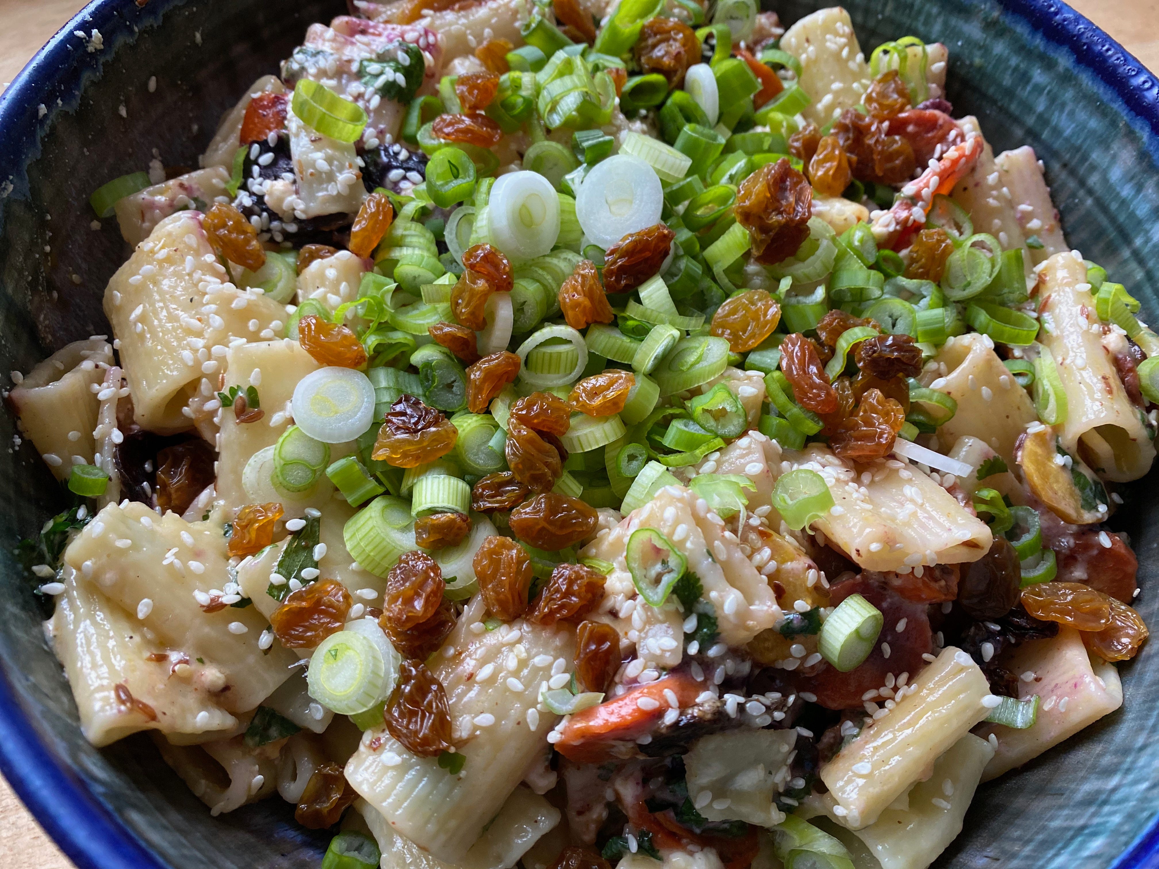 Closeup of a blue ceramic bowl filled with pasta, roasted carrots, sliced scallions, sesame seeds, and golden raisins.