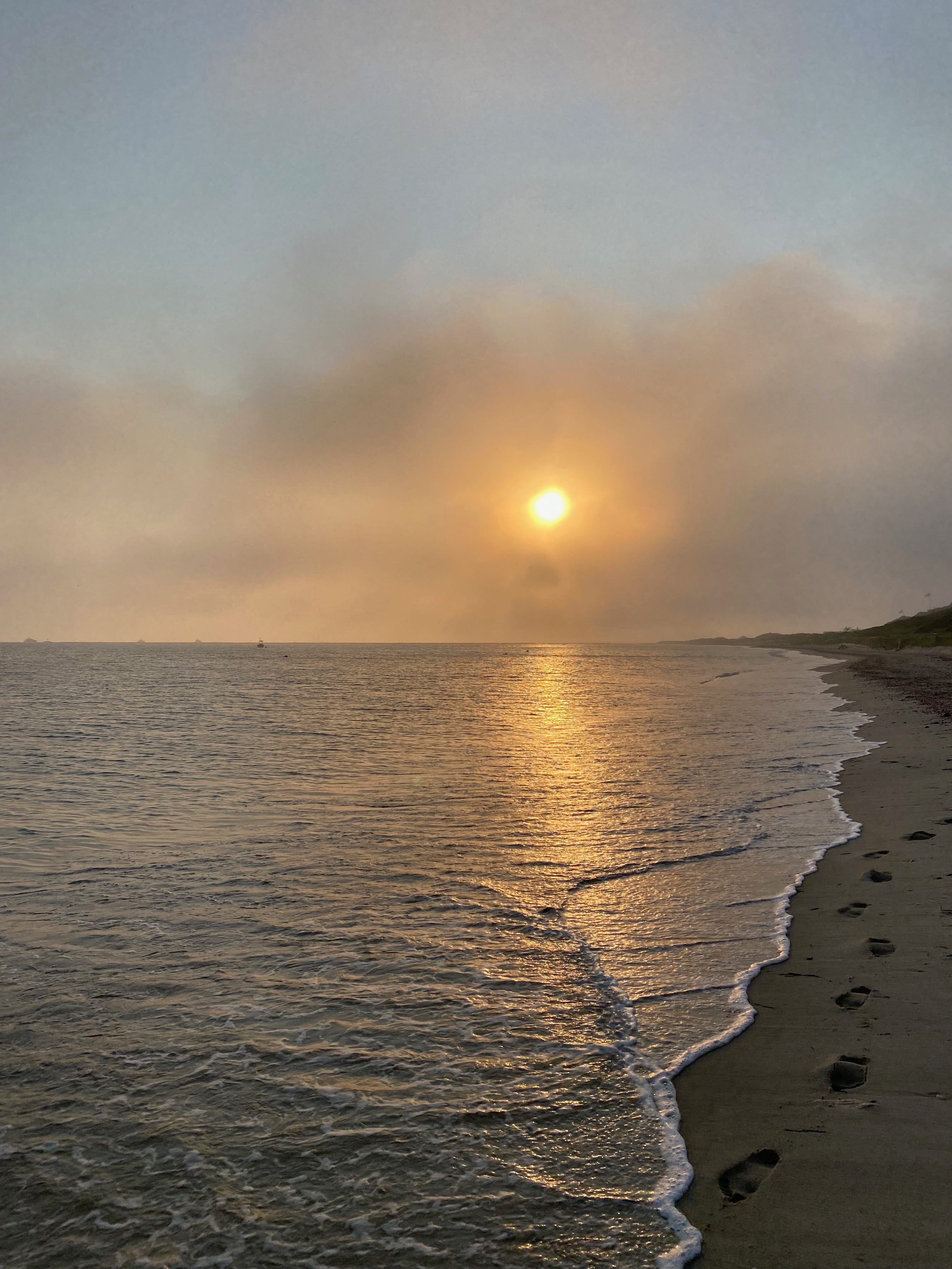 Sunrise over the ocean. The sun is bright gold, low on the horizon, and surrounded by dark clouds. Its light spills across the water. A wave is breaking and a thin strip of beach is visible.