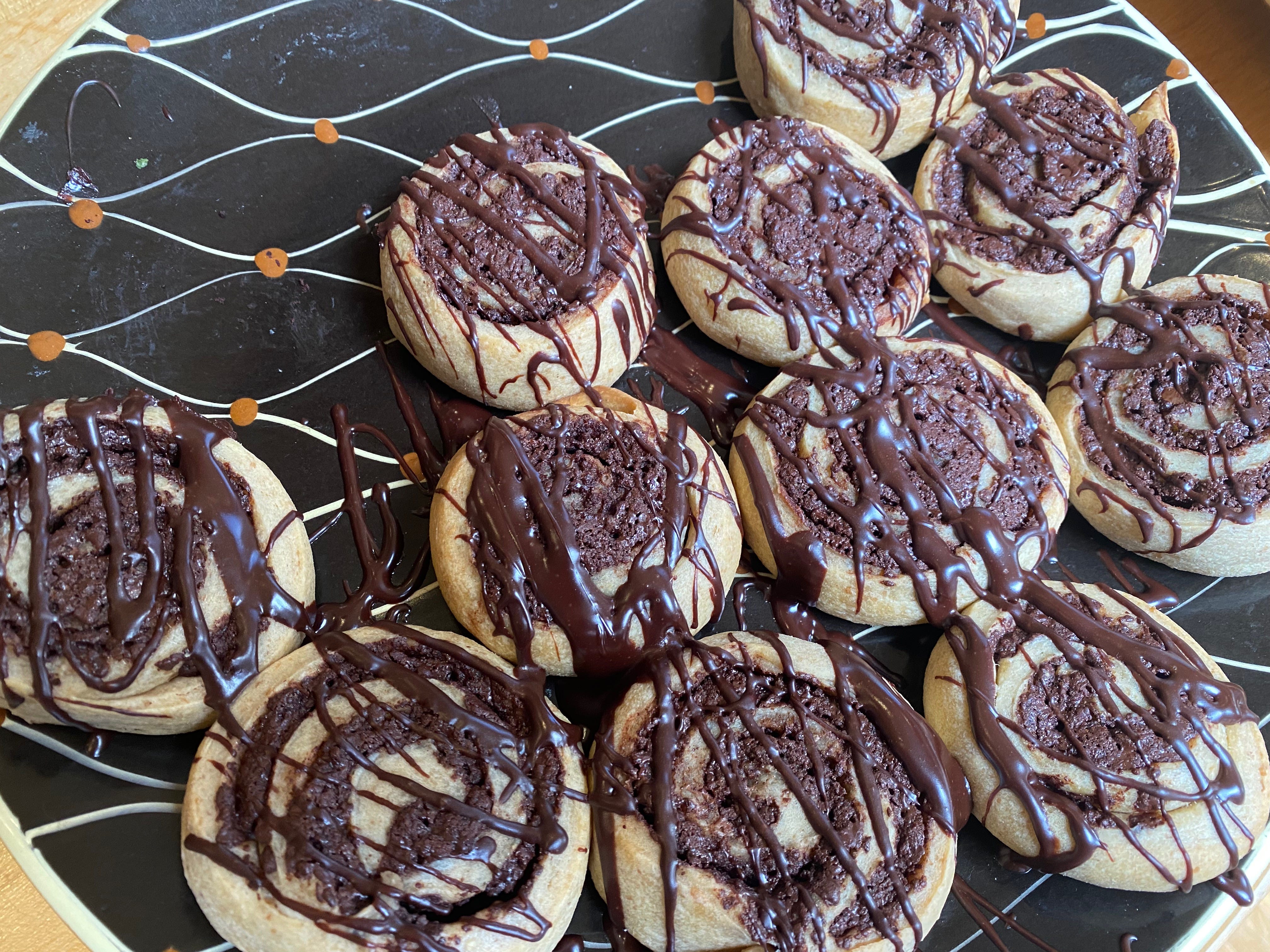 A black serving plate holds several round chocolate swirl buns, drizzled with chocolate ganache.
