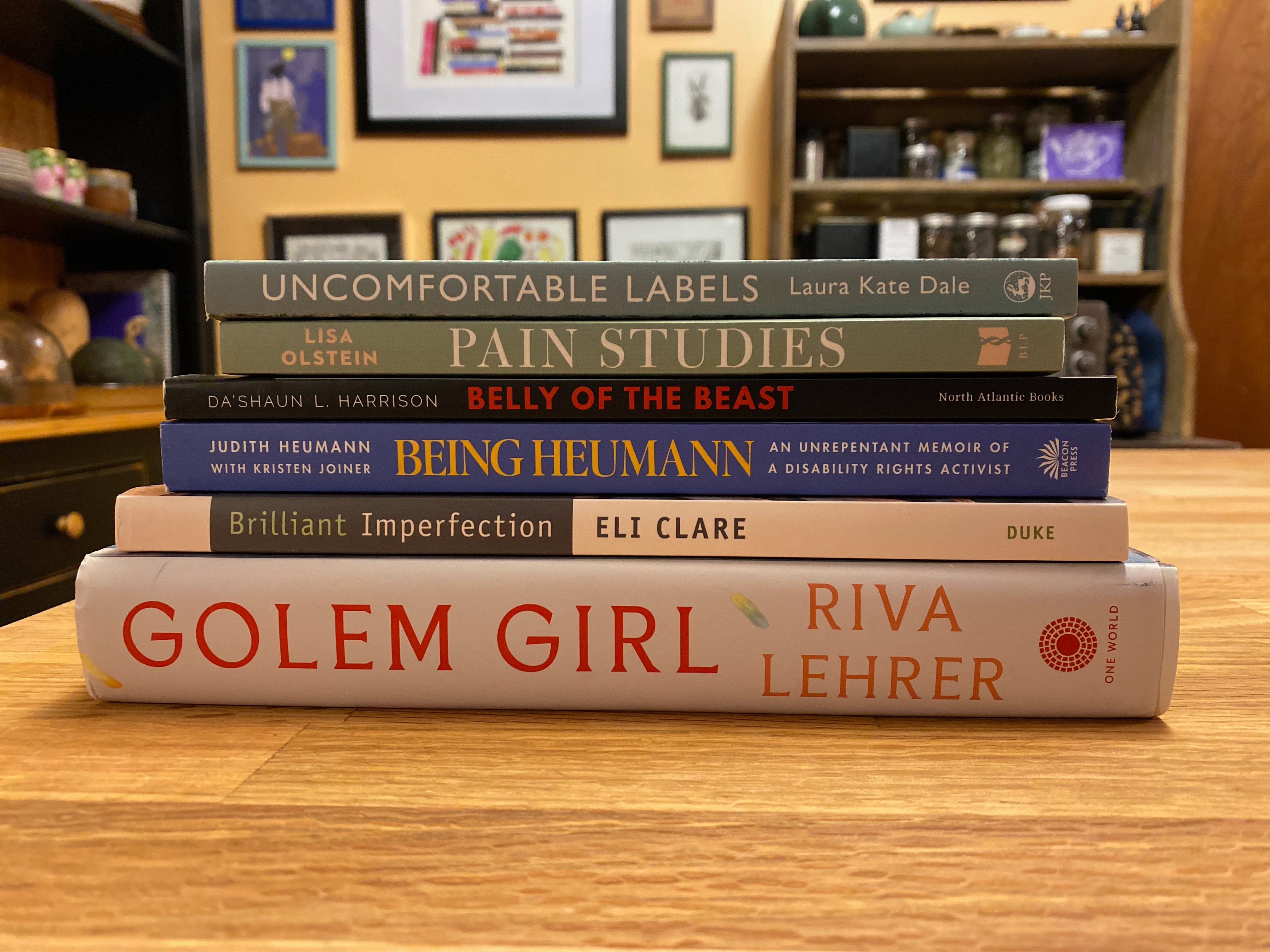 A stack of books by disabled authors on a kitchen island. In the background, slightly blurred: a shelf full of tins of tea, part of a hutch holding ceramics, and a yellow wall filled with colorful prints.