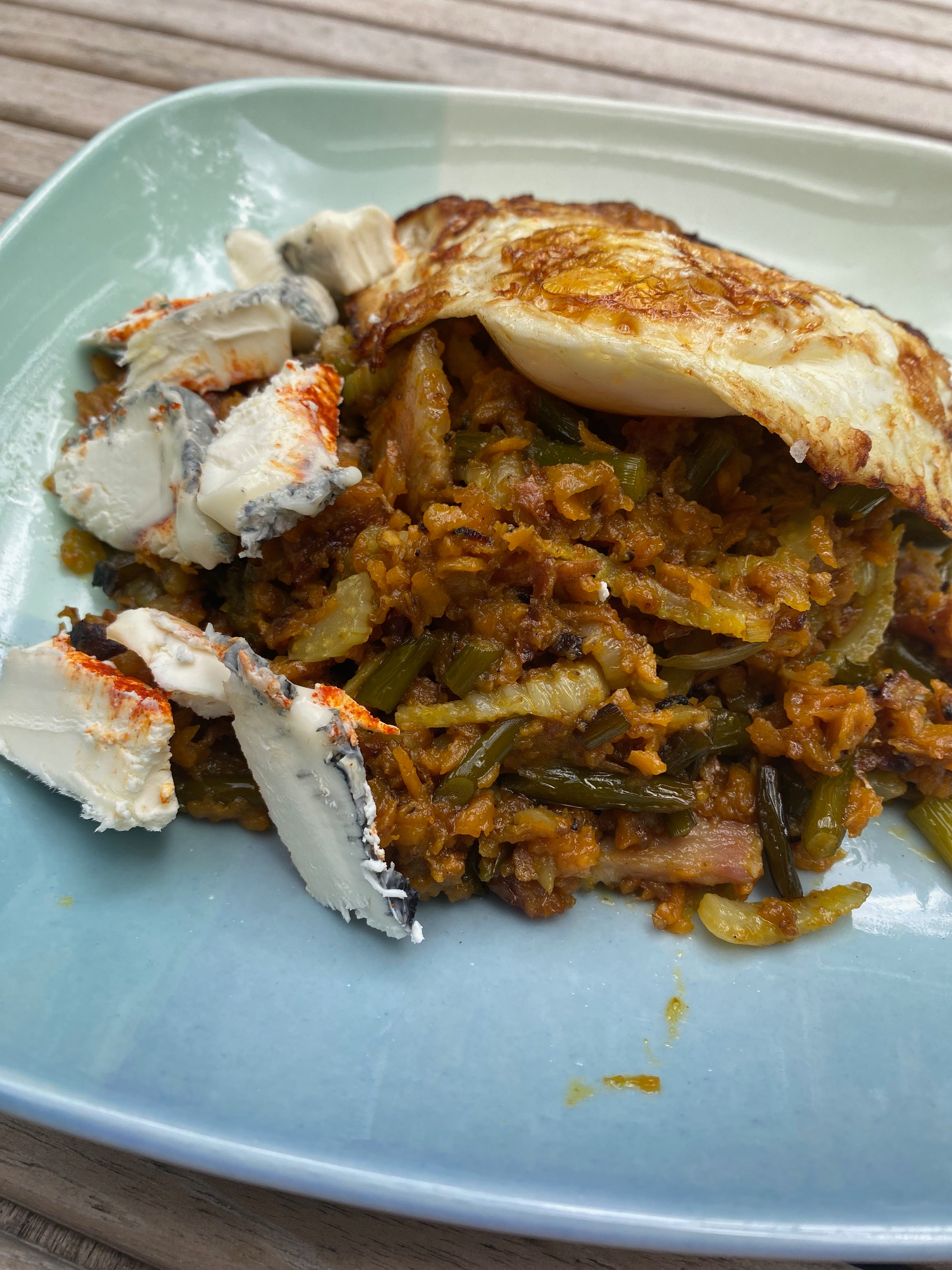 A fried egg and slices of thick, gooey soft cheese coated in ash sit on top of a pile of sweet potato hash on a ceramic plate.