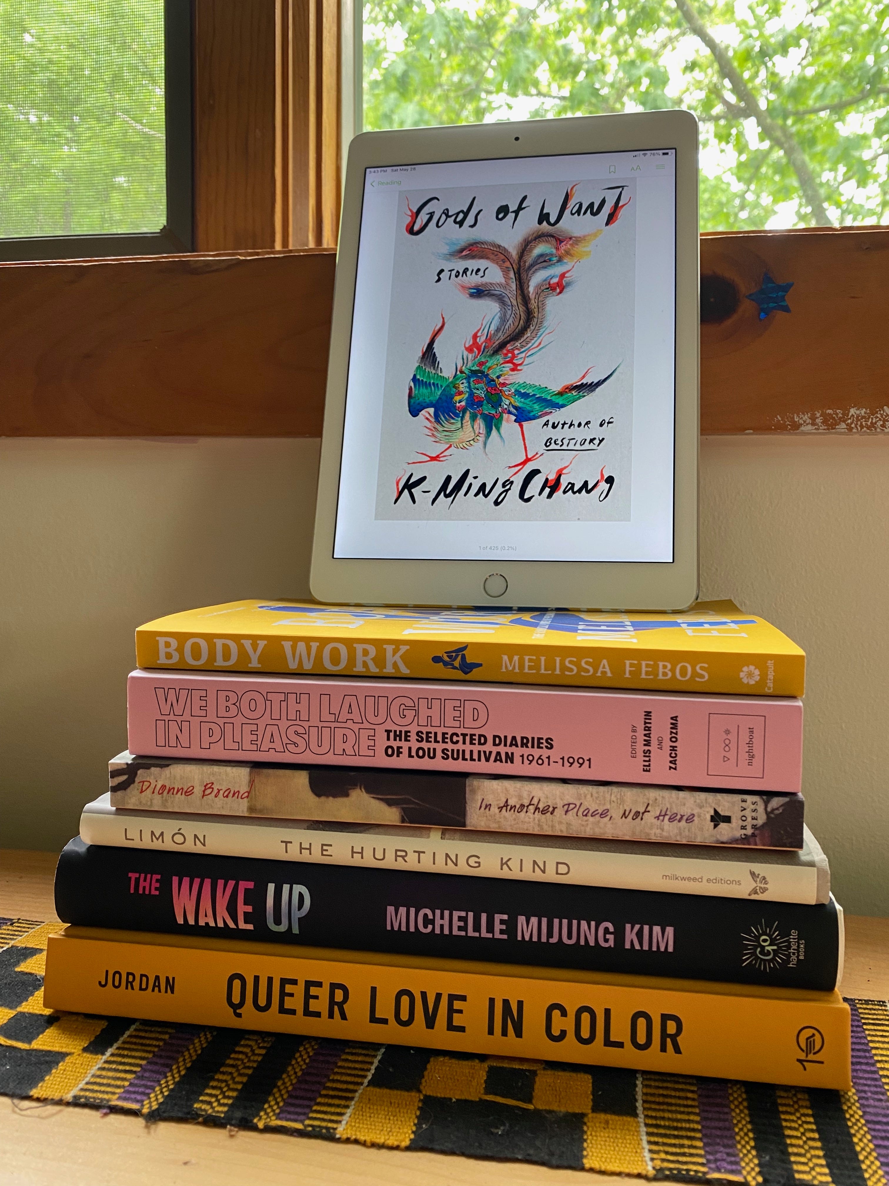 The books listed stacked on a bookshelf under a sunny window. Gods of Want is displayed on an iPad propped on top of the stack.