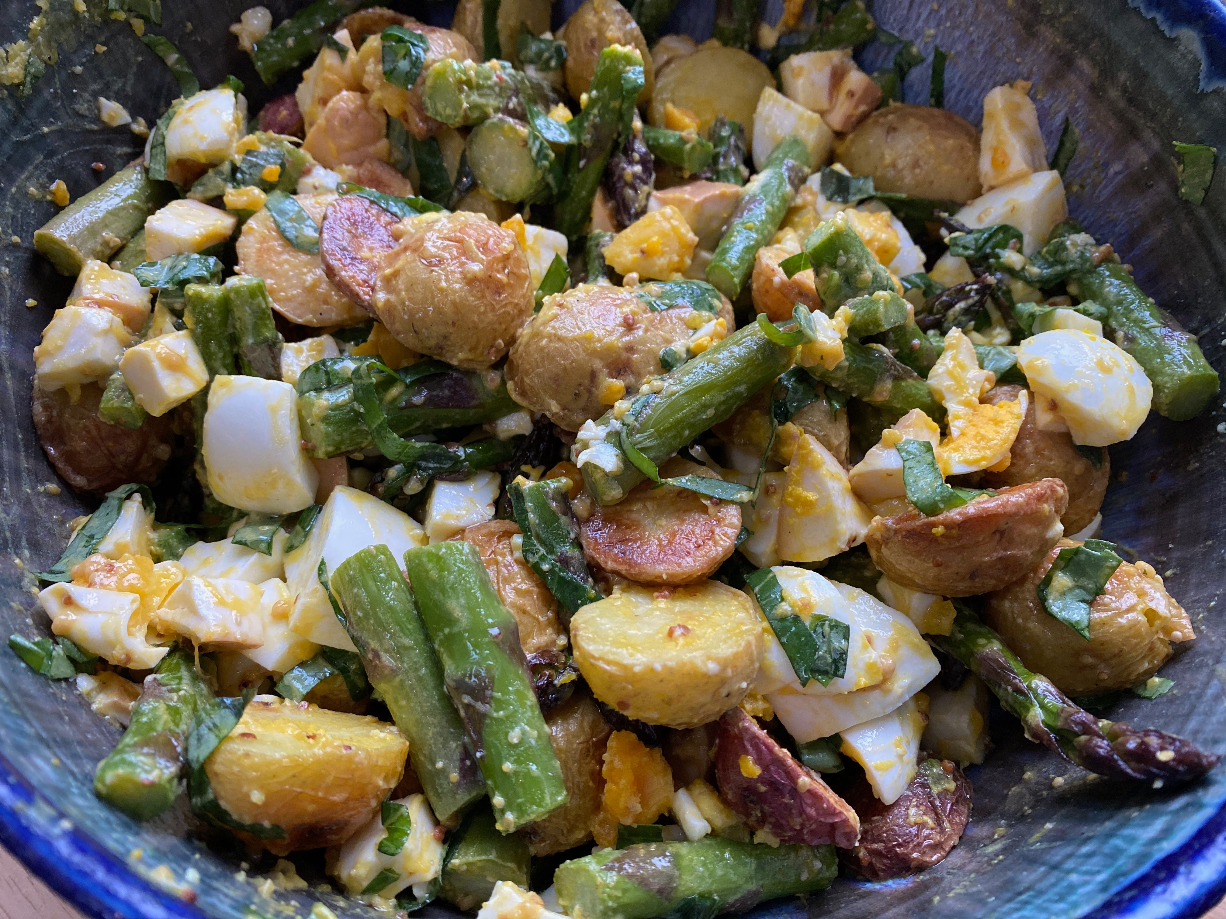 A closeup of a blue ceramic bowl: roasted potatoes, small asparagus spears, chopped hardboiled eggs, and strips of basil are all coated in a mustardy dressing.