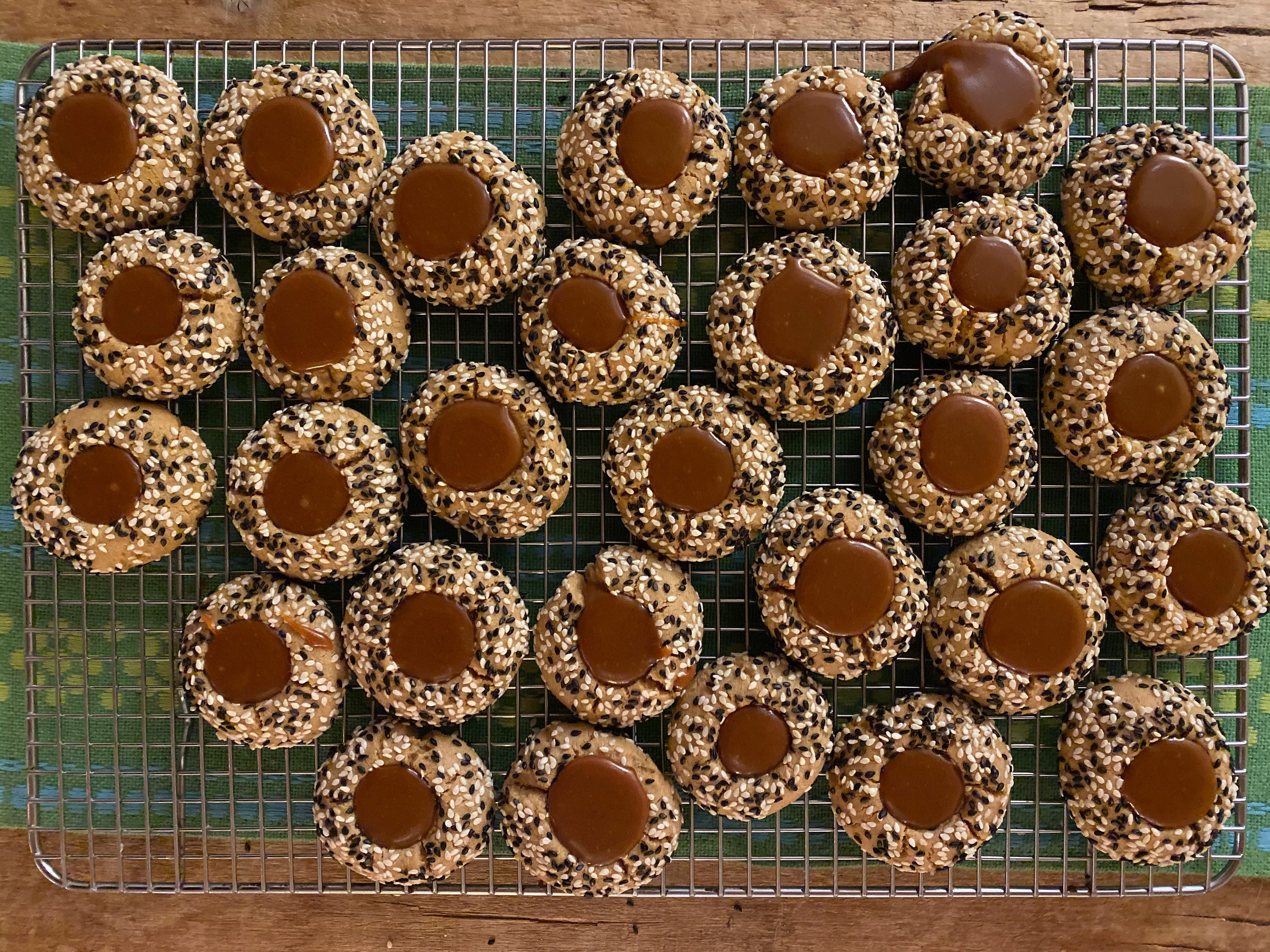 Many round cookies, filled with dark brown caramel and studded with white and black sesame seeds, sit on a wire cooling rack on wooden table covered in a green cloth.