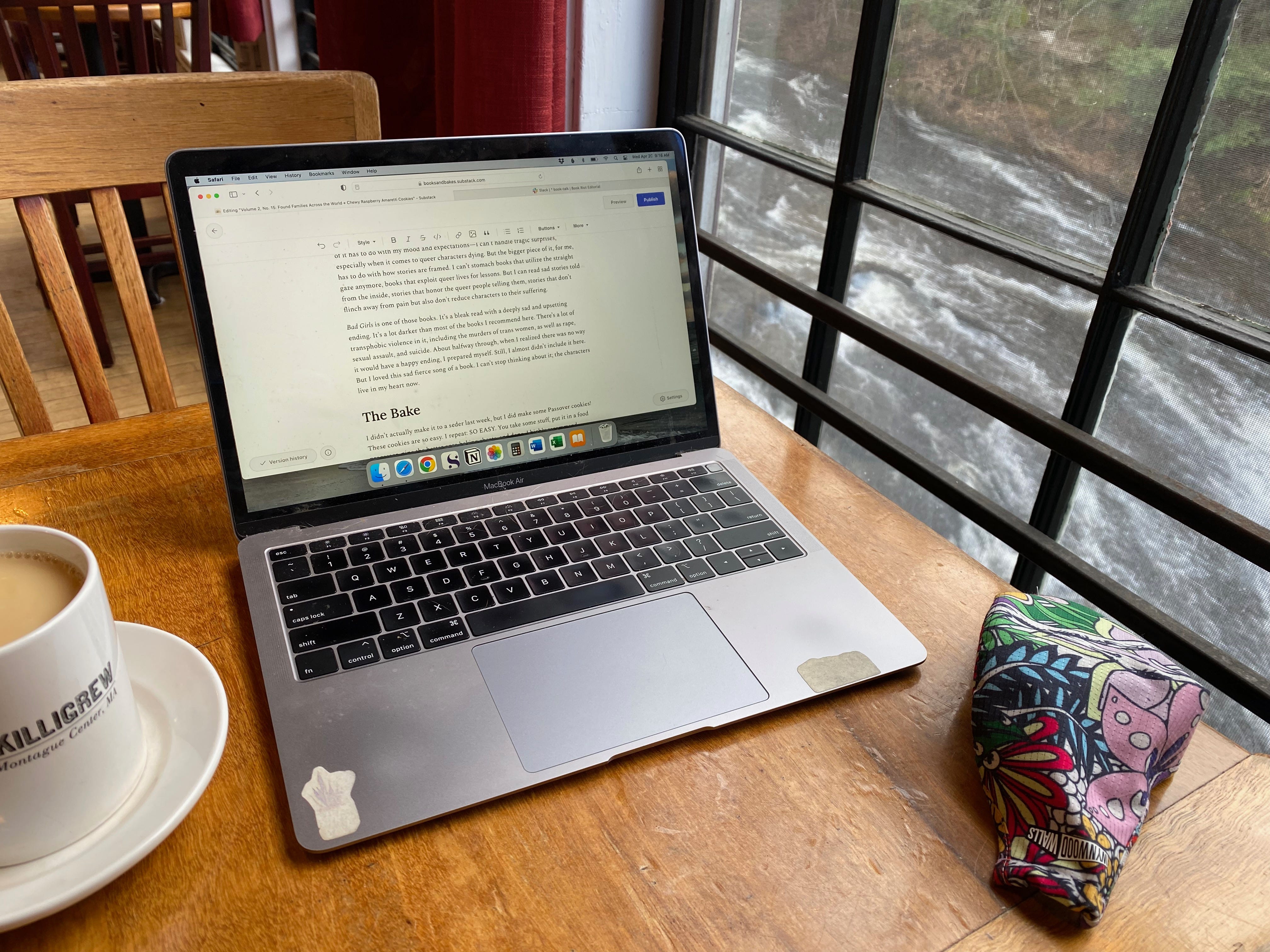 A laptop sits open a wooden cafe table next to a mug of tea and a colorful mask. Outside the window is a rushing river.