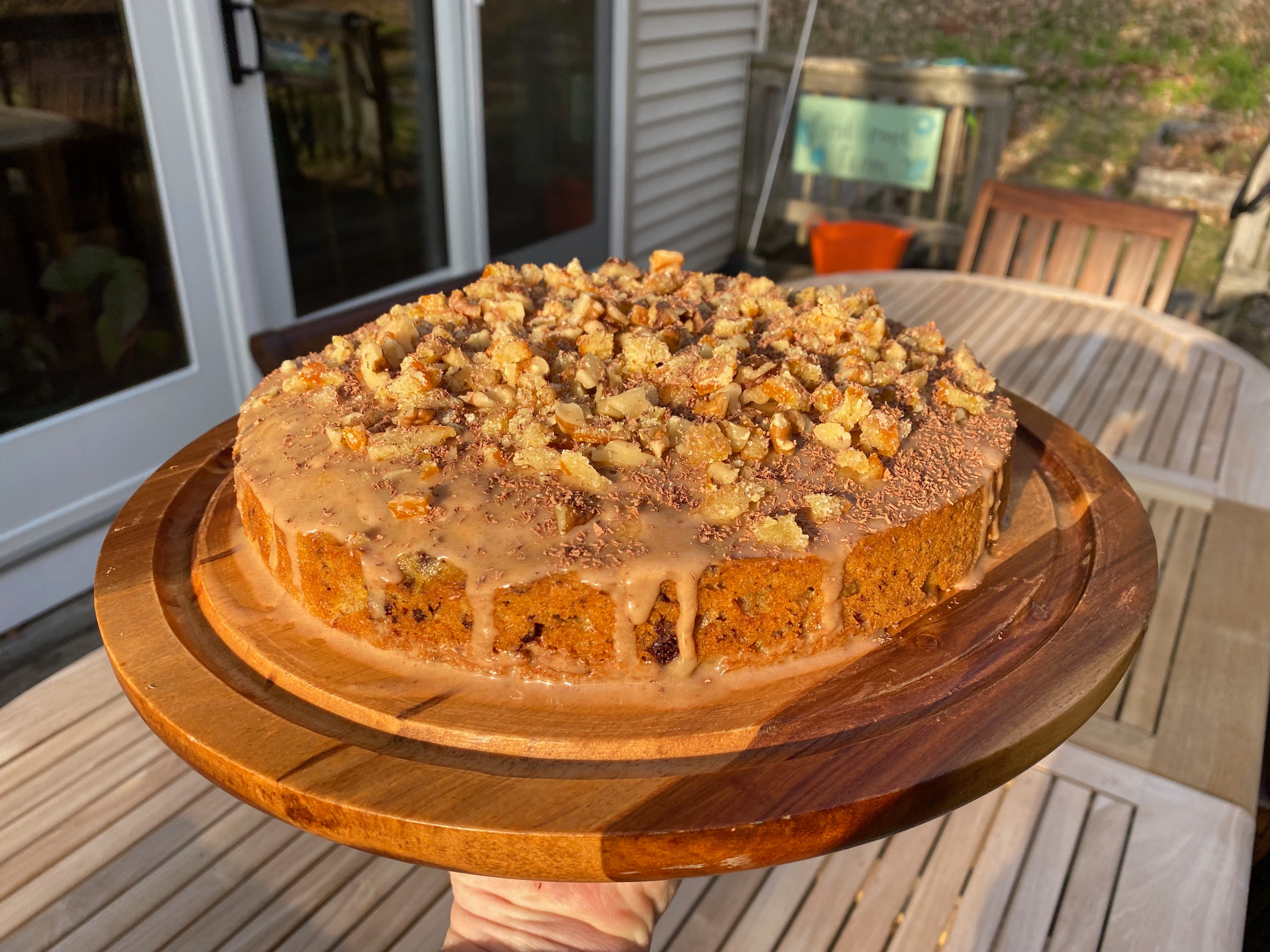 My hand holding a wooden cake platter in the afternoon sunlight in front of an outdoor table and the porch sliders. The round cake is topped with chopped walnuts, candied orange, and shaved chocolate, with glaze dripping down the side.