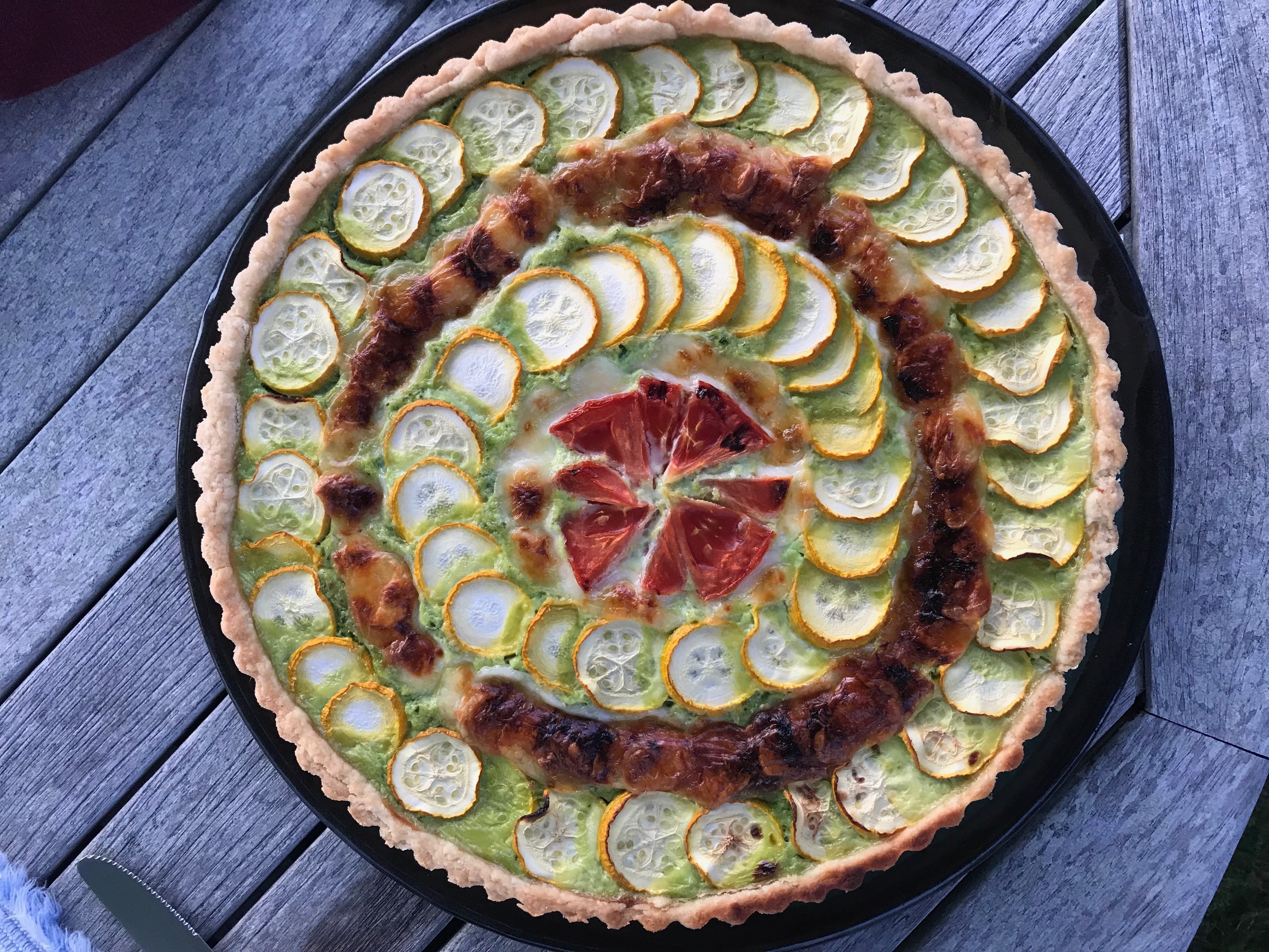 A round fluted tart on a wooden table. The green garlic scape filling is topped with concentric circles of zucchini, summer squash, sun-dried tomatoes, and browned mozzarella cheese.