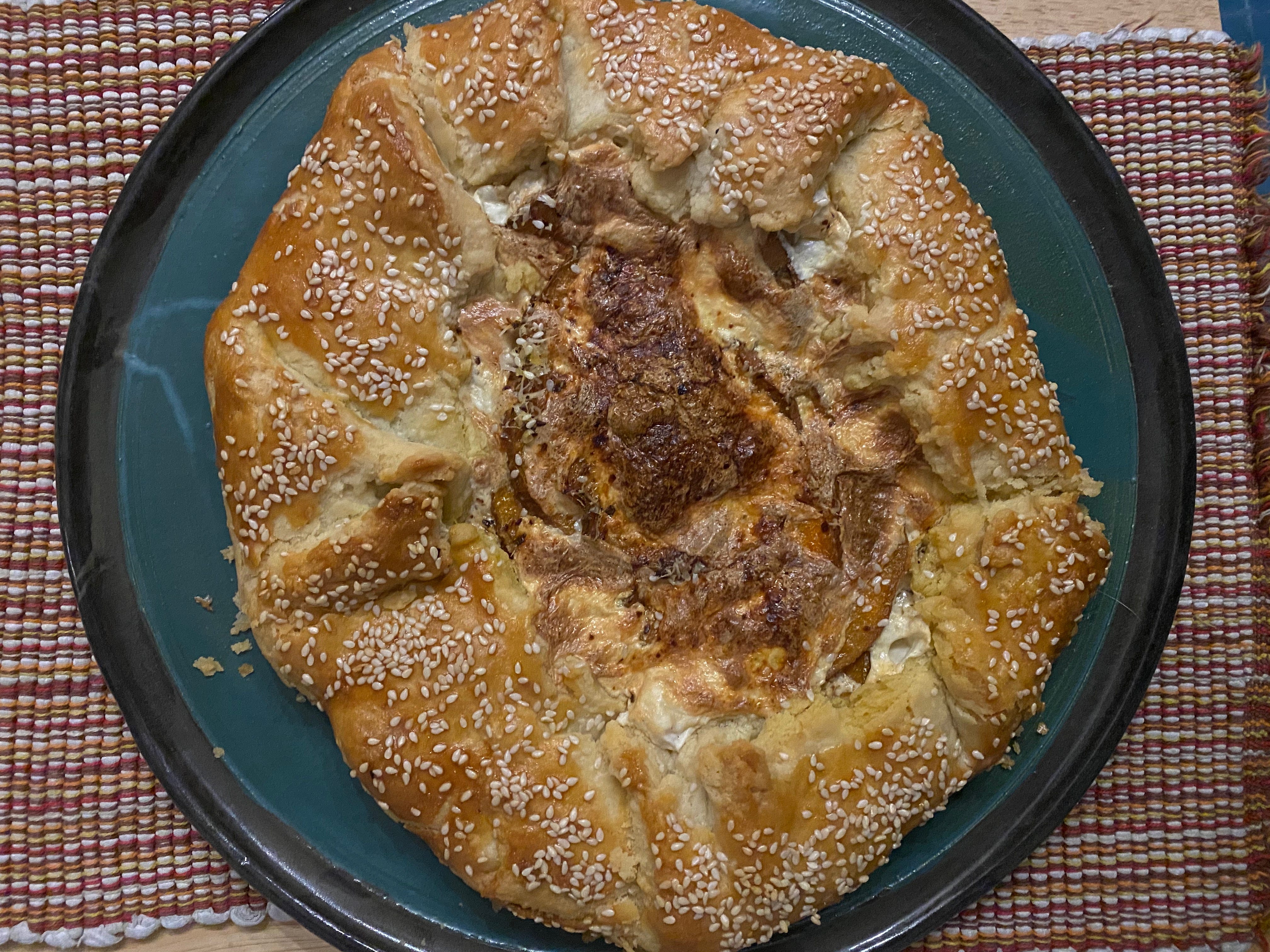 A galette sits on a blue ceramic plate. It’s roundish, with golden brown crust scattered with sesame seeds. The filling in the center is browned, bubbly mozzarella cheese.