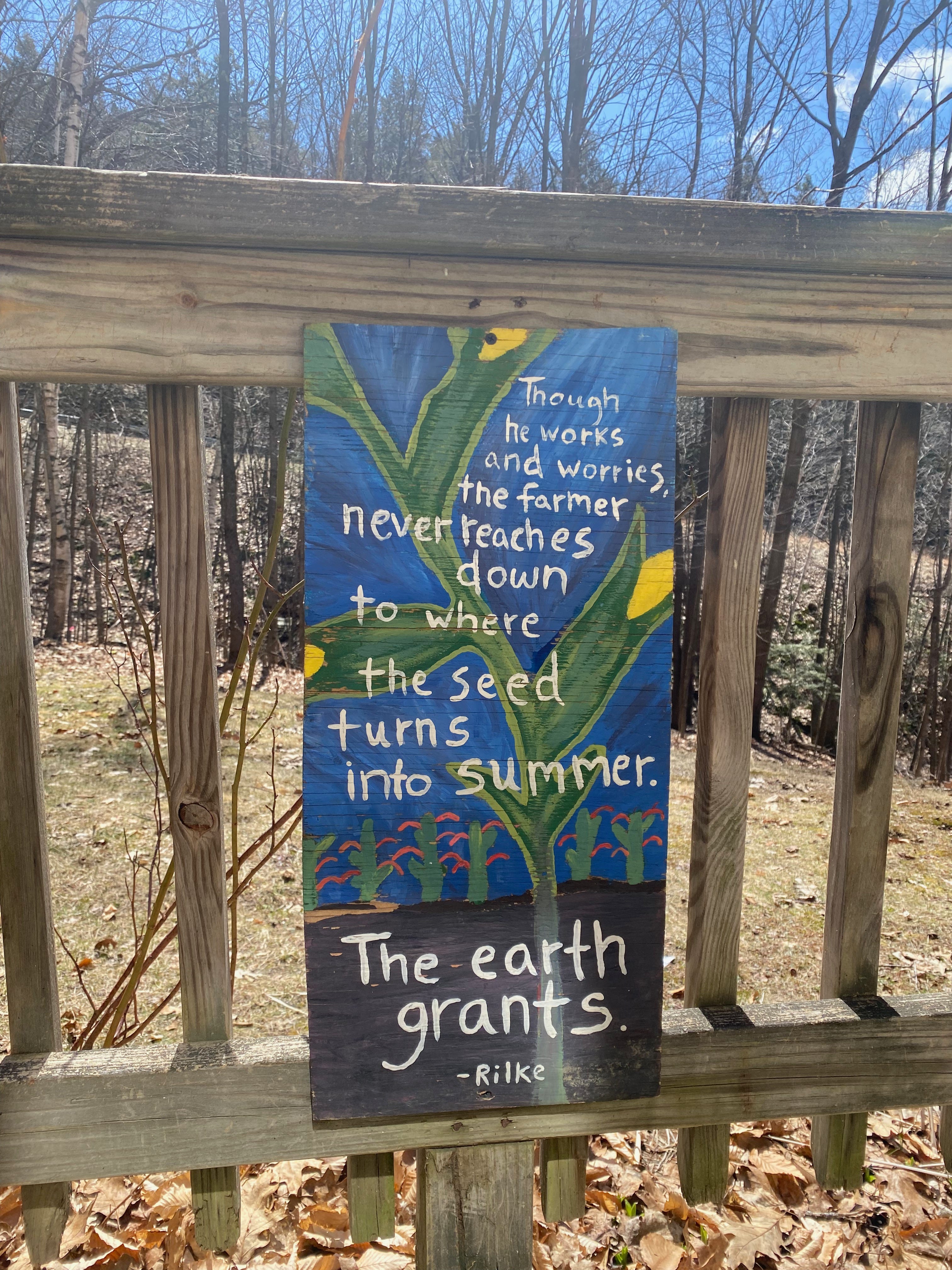 A handprinted wooden sign hangs on a wooden porch railing in front of blue sky and bare spring trees. The sign is painted blue, with a large corn stalk reaching up into the sky, its roots extending into the dirt below. Behind the corn is a small row of stylized plants. The quote painted in white on top of the image reads: “Though he works and worries, the farmer never reaches down to where the seed turns into summer. The earth grants.” (Rilke)