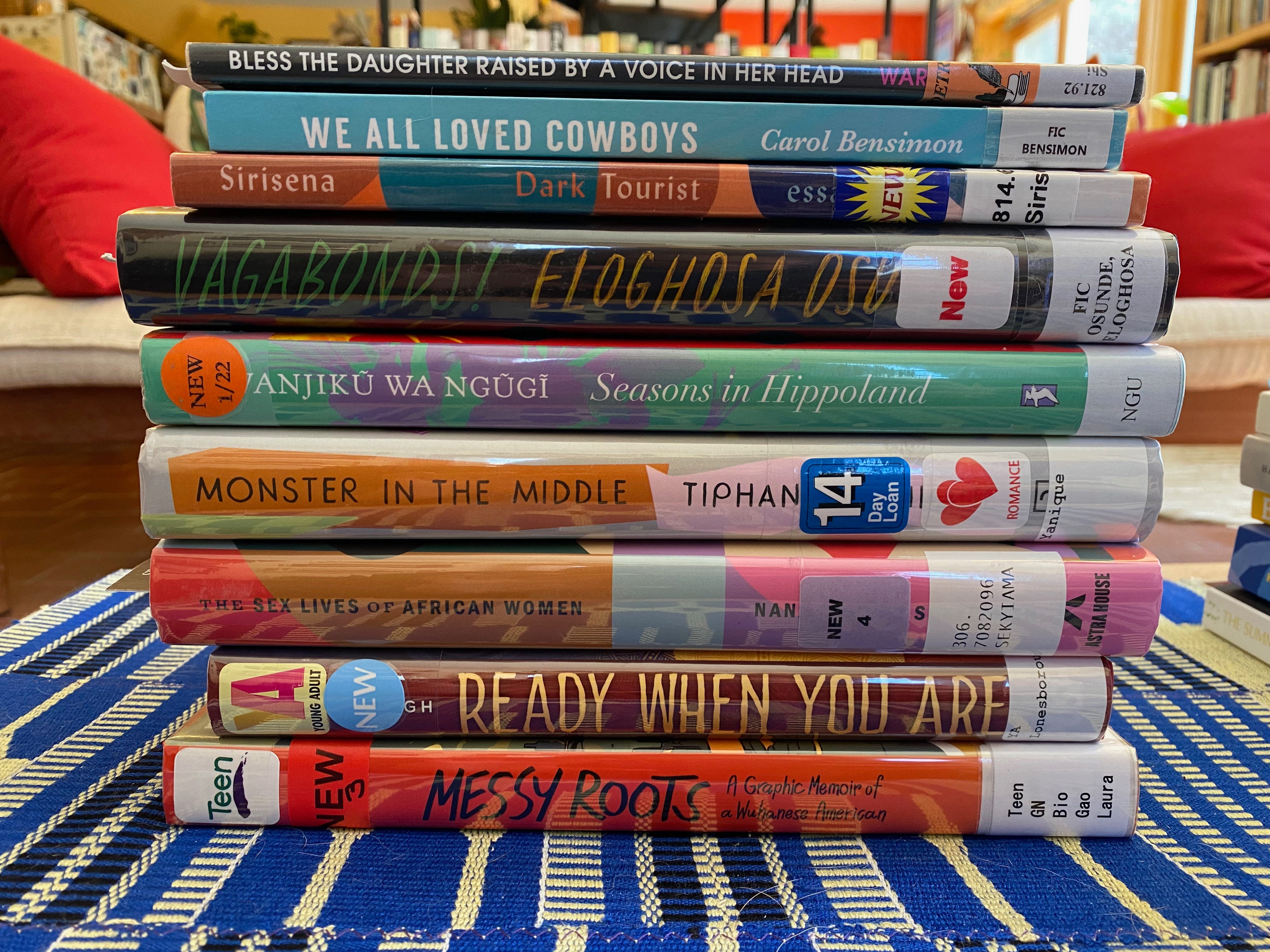 A stack of library books on a coffee table in a sunny living room. Books include Messy Roots, Ready When You Are, The Sex Lives of African Women, Monster in the Middle, Seasons in Hippoland, Vagabonds!, Dark Tourist, We All Loved Cowboys, and Bless the Daughter Raised by a Voice in her Head.