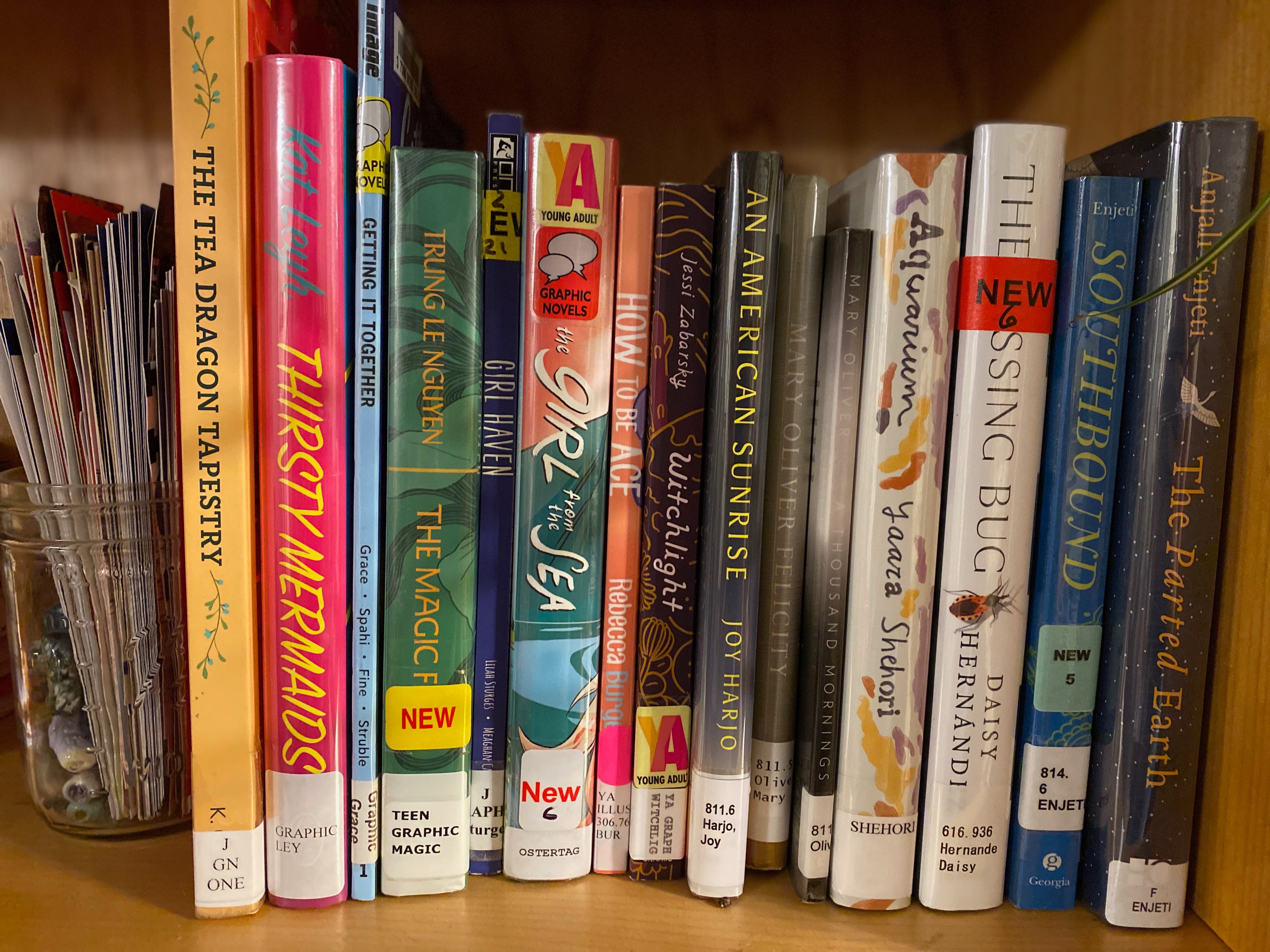A shelf of library books bookended on one side by a jar full of bookmarks. Books are: The Tea Dragon Tapestry, Thirsty Mermaids, Getting It Together, The Magic Fish, Girl Haven, The Girl from the Sea, How to be Ace, Witchlight, An American Sunrise, Felicity, A Thousand Mornings, Aquarium, The Kissing Bug, Southbound, The Parted Earth.