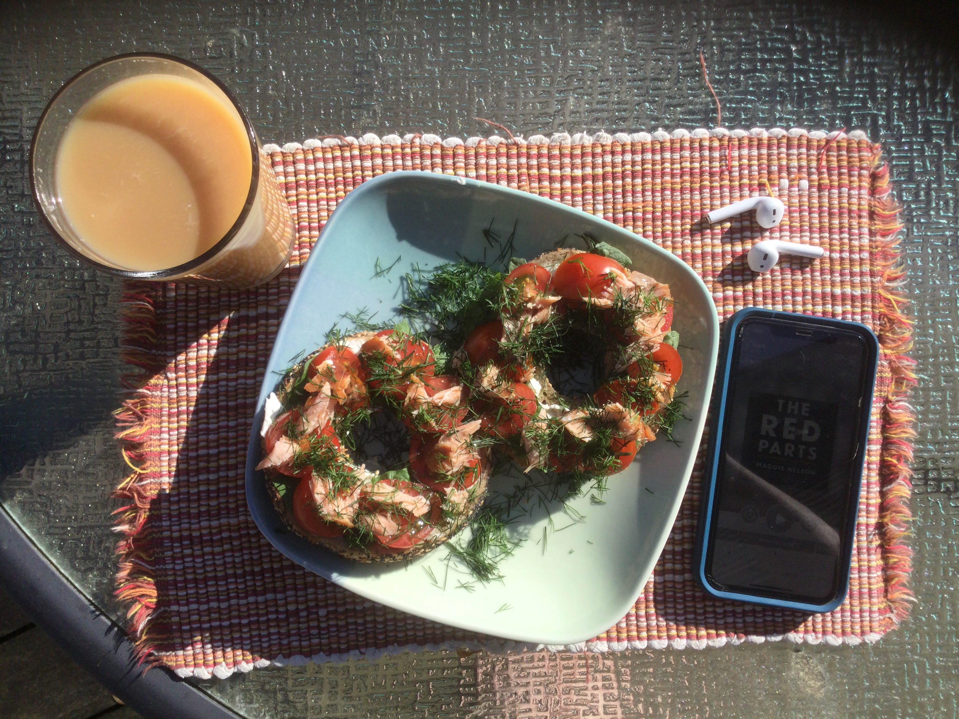 View from above of a square blue and green plate sitting on a woven placemat. There’s a tall glass of iced tea on the left and a phone and a pair of earbuds on the left. On the plate are two bagel halves, topped with tomatoes, smoked salmon, and lots of dill.