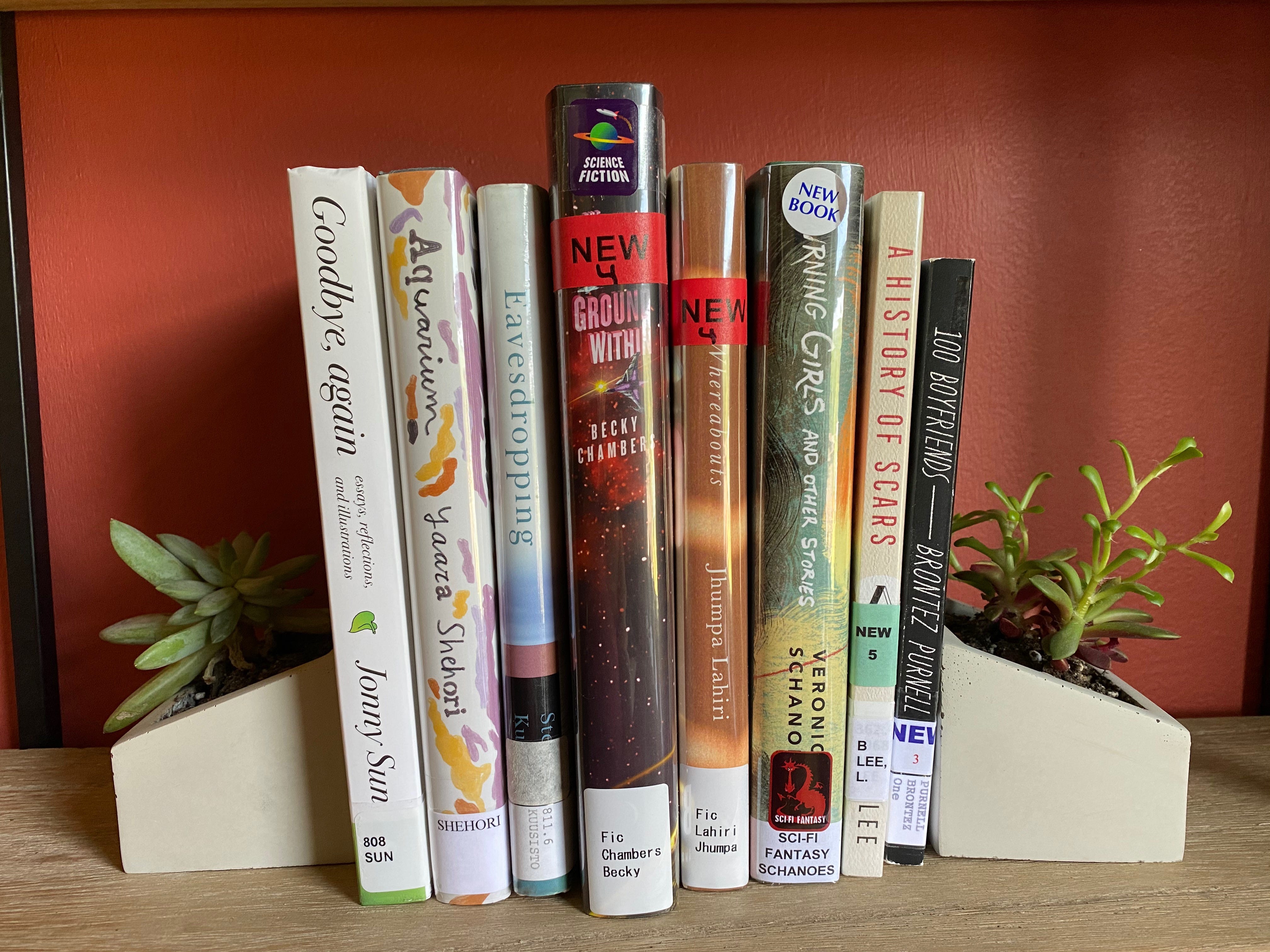 A shelf of library books between succulent planter bookends. The books are: Goodbye, Again, Aquarium, Eavesdropping, The Galaxy, and the Ground Within, Whereabouts, Burning Girls and Other Stories, A History of Scars, and 100 Boyfriends.