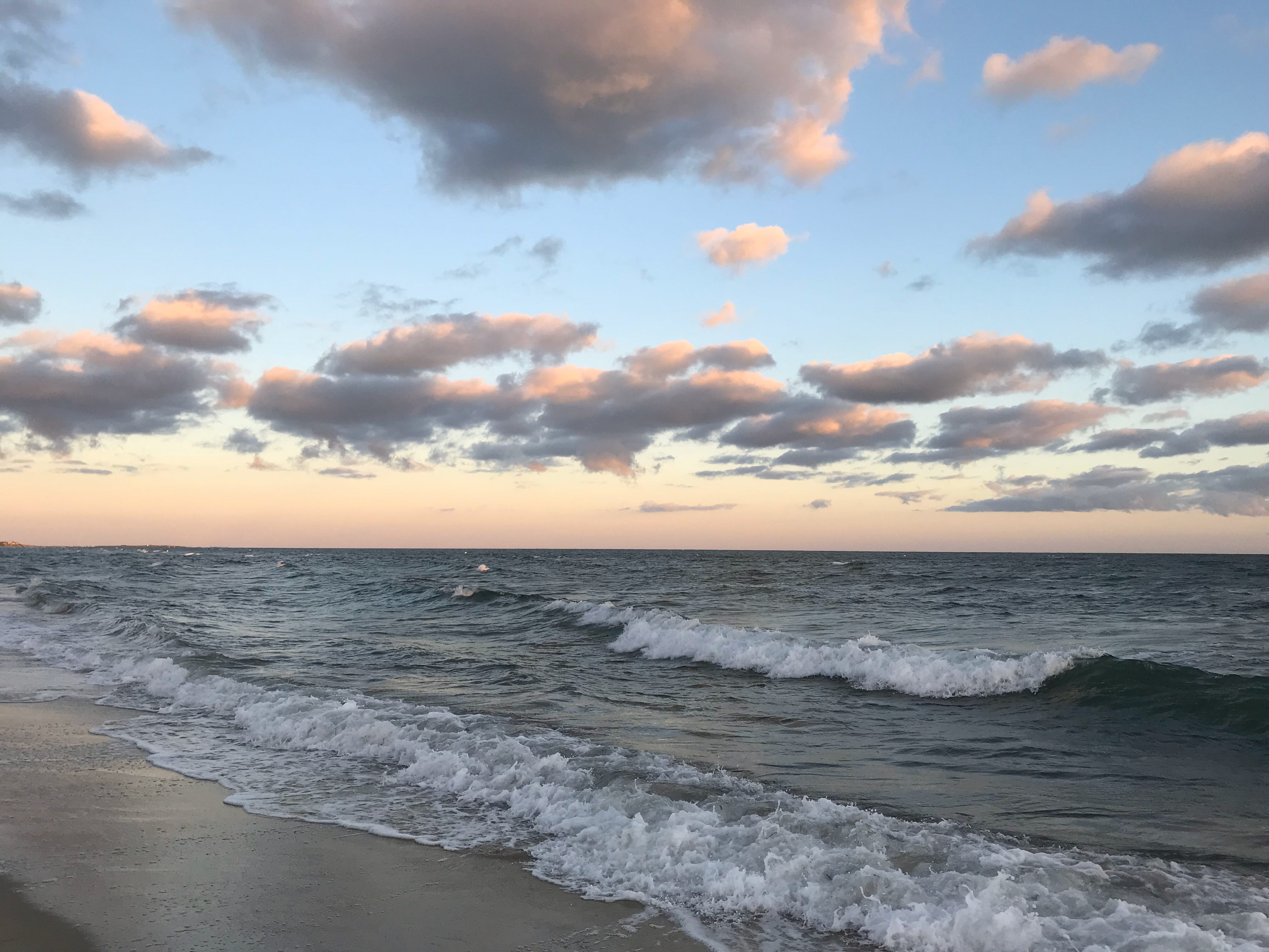 A view of the dark blue ocean under a pale blue sky, dotted with pink and blue clouds. Waves are breaking along the beach.