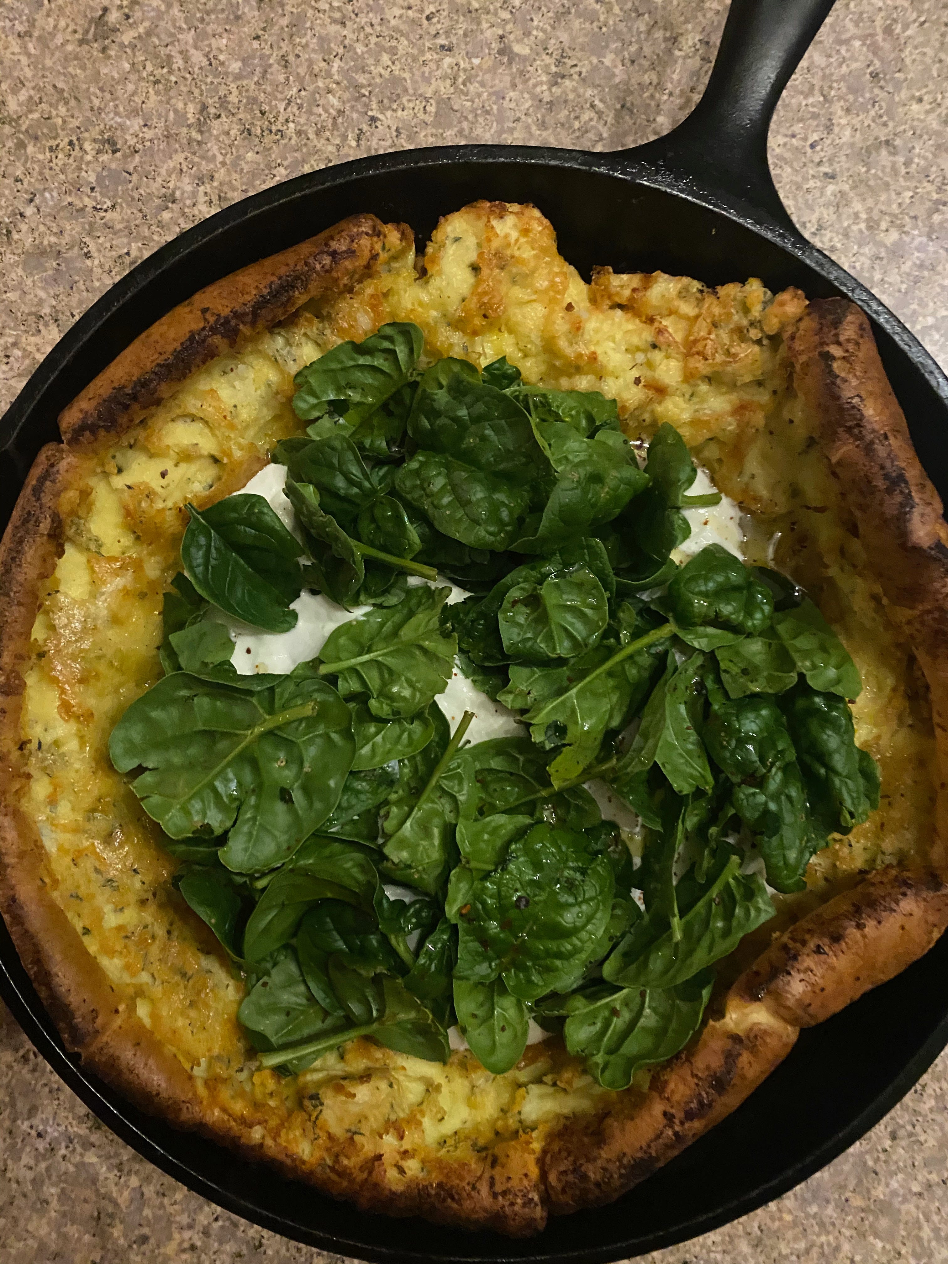 A cast iron pan, viewed from above, holding a Dutch baby. It’s puffed and golden, with darker brown edges. The center is filled with ricotta, which you can just see beneath a pile of spinach leaves.