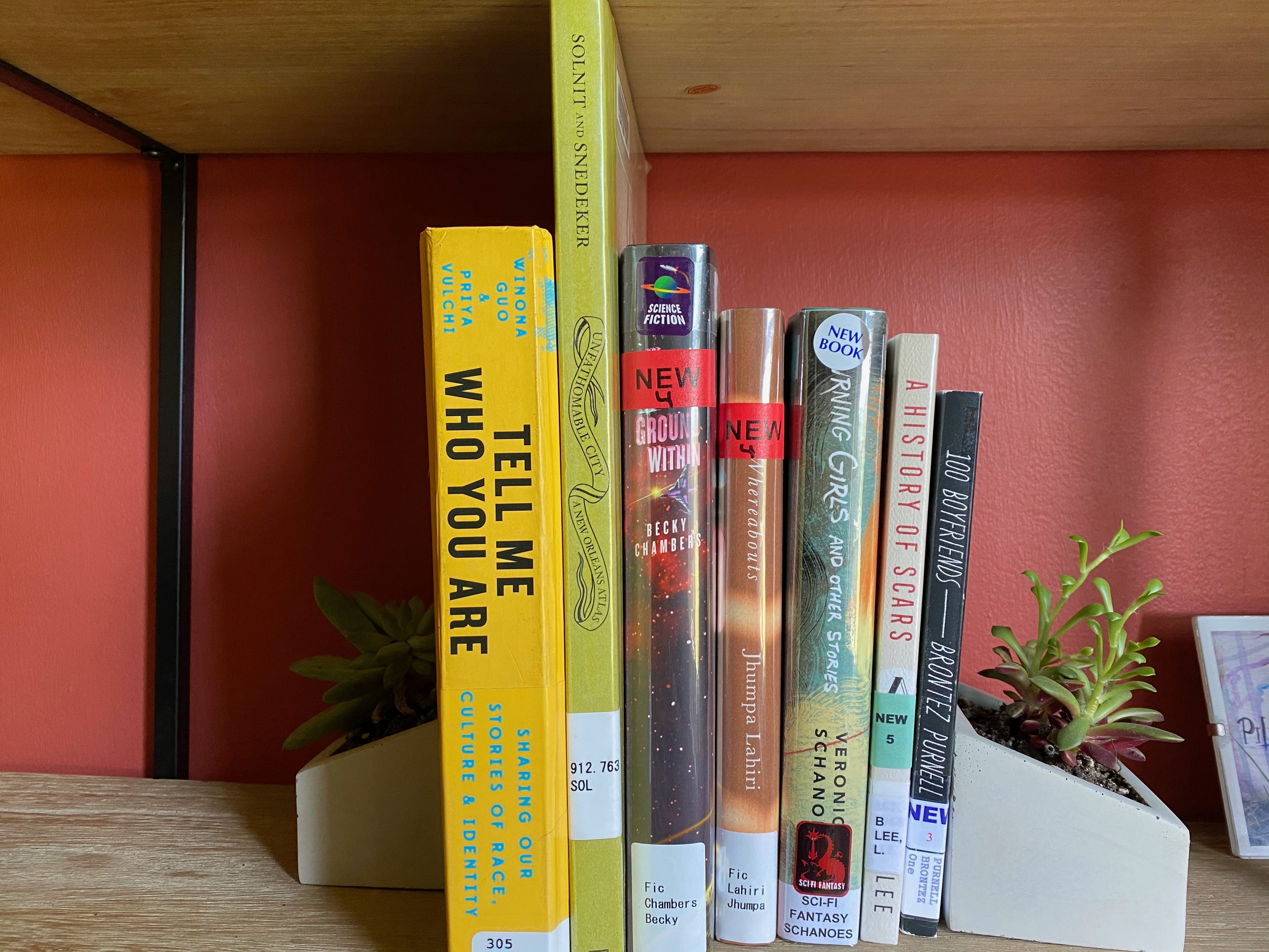 A shelf of library books between two succulent planter bookends. The books are:&nbsp;Tell Me Who You Are, Unfathomable City, The Galaxy, and the Ground Within, Whereabouts, Burning Girls, A History of Scars, and 100 Boyfriends.
