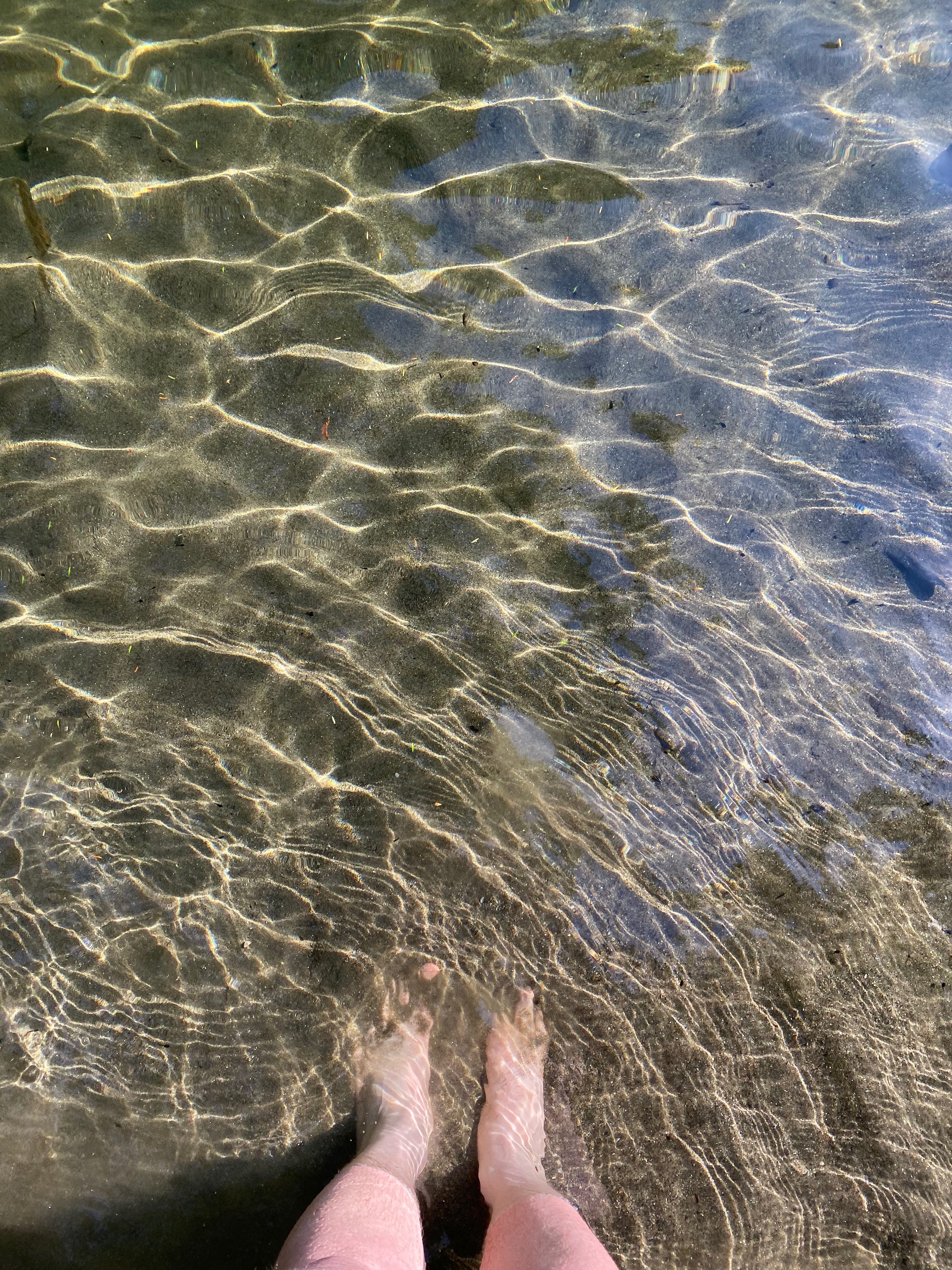 A view, from above, of my bare feet in a pool of clear water. The surface of the water is dappled with sunlight, making ripples and lines of different shapes.