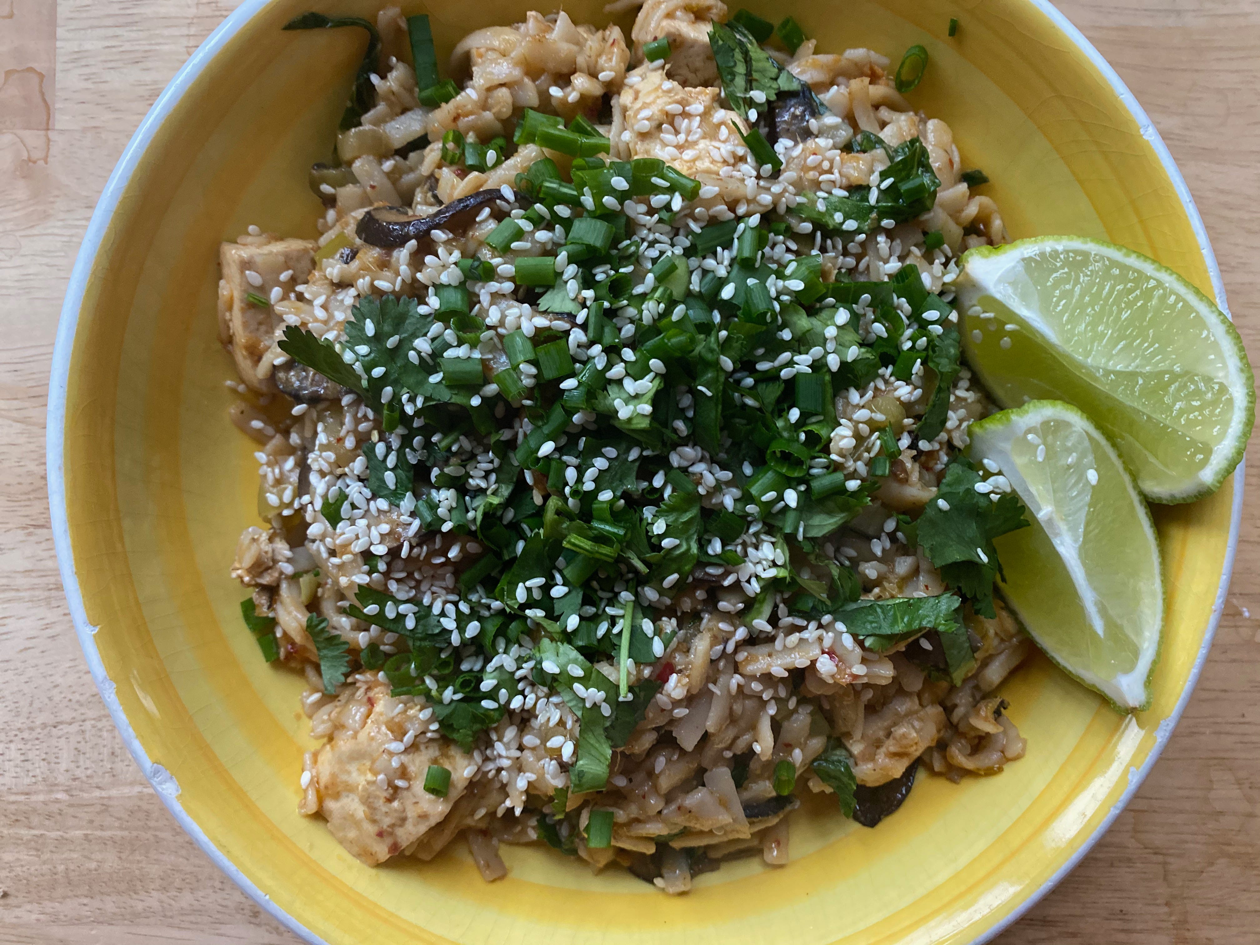 A shallow, bright yellow bowl full of red curry noodles. There’s a pig pile of cilantro, scallions, and sesame seeds on top of the noodles, and two lime wedges on one side.