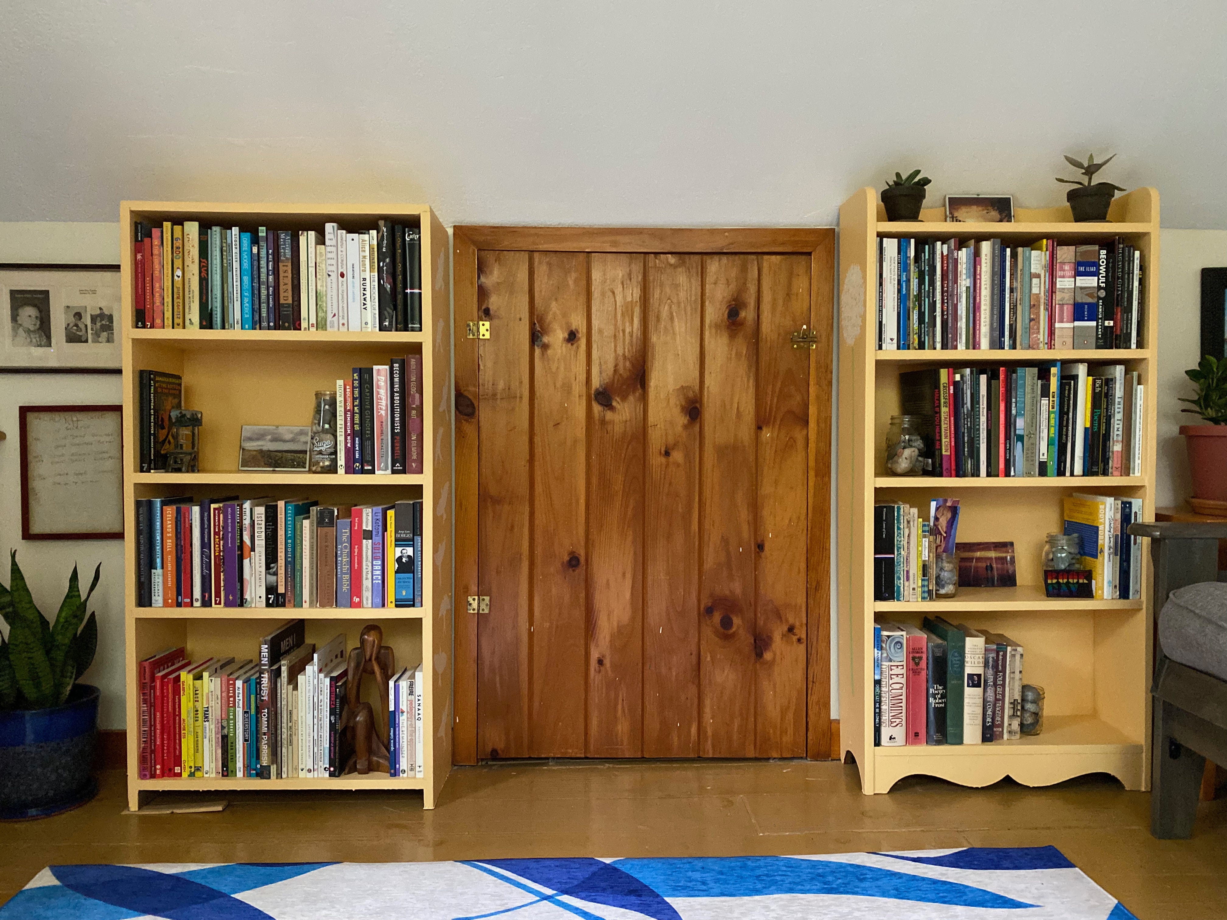 Two small orange bookshelves, both full of books, plants, and photos, stand against the wall on either side of a small closet door.