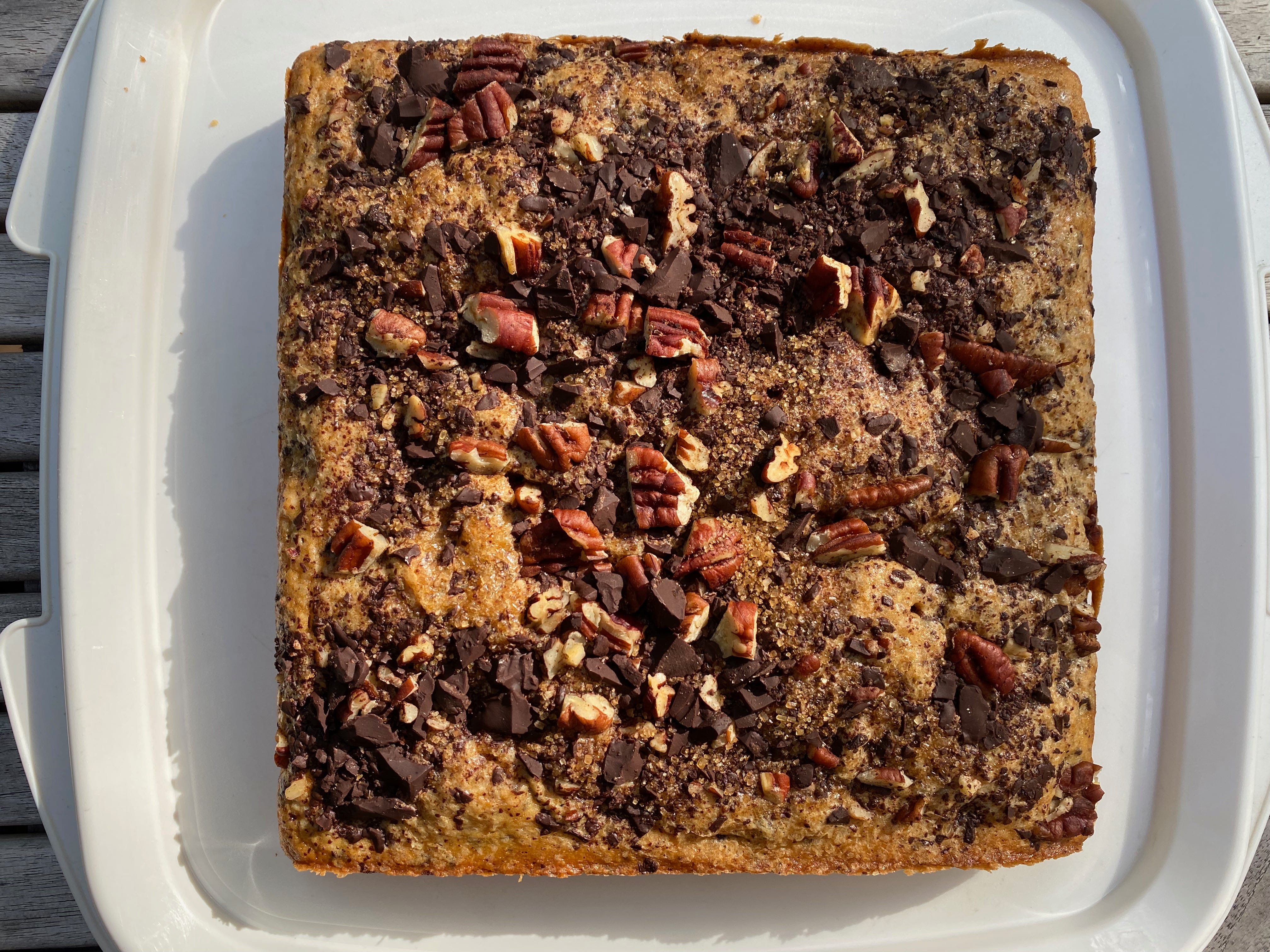 A square cake topped with chopped chocolate and pecans sits on a white cake tray.