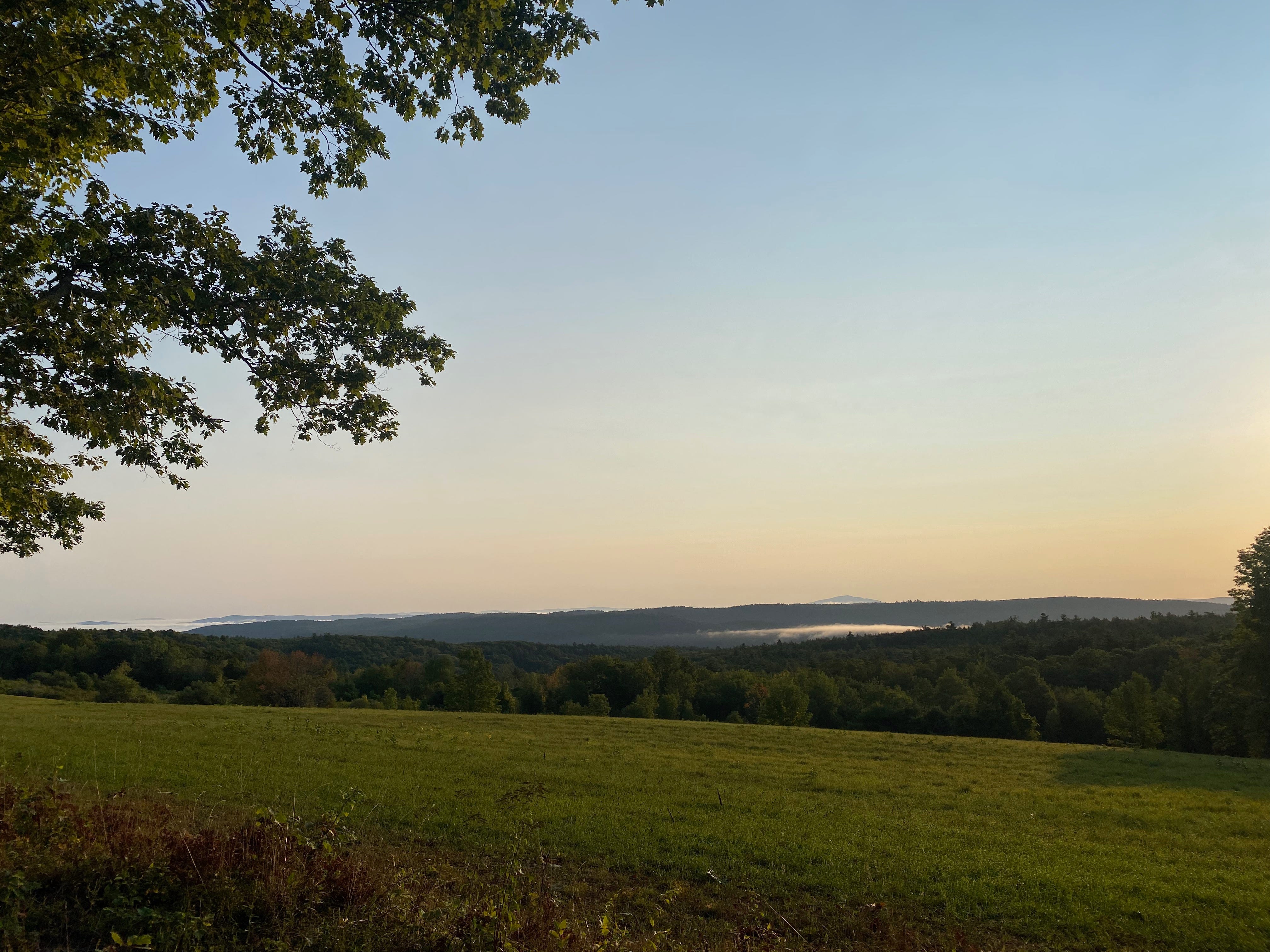 A green field at the top of the hill, in front of a vista of distant mountains and hills, partially covered in clouds. The sky is pale gold, fading into blue.