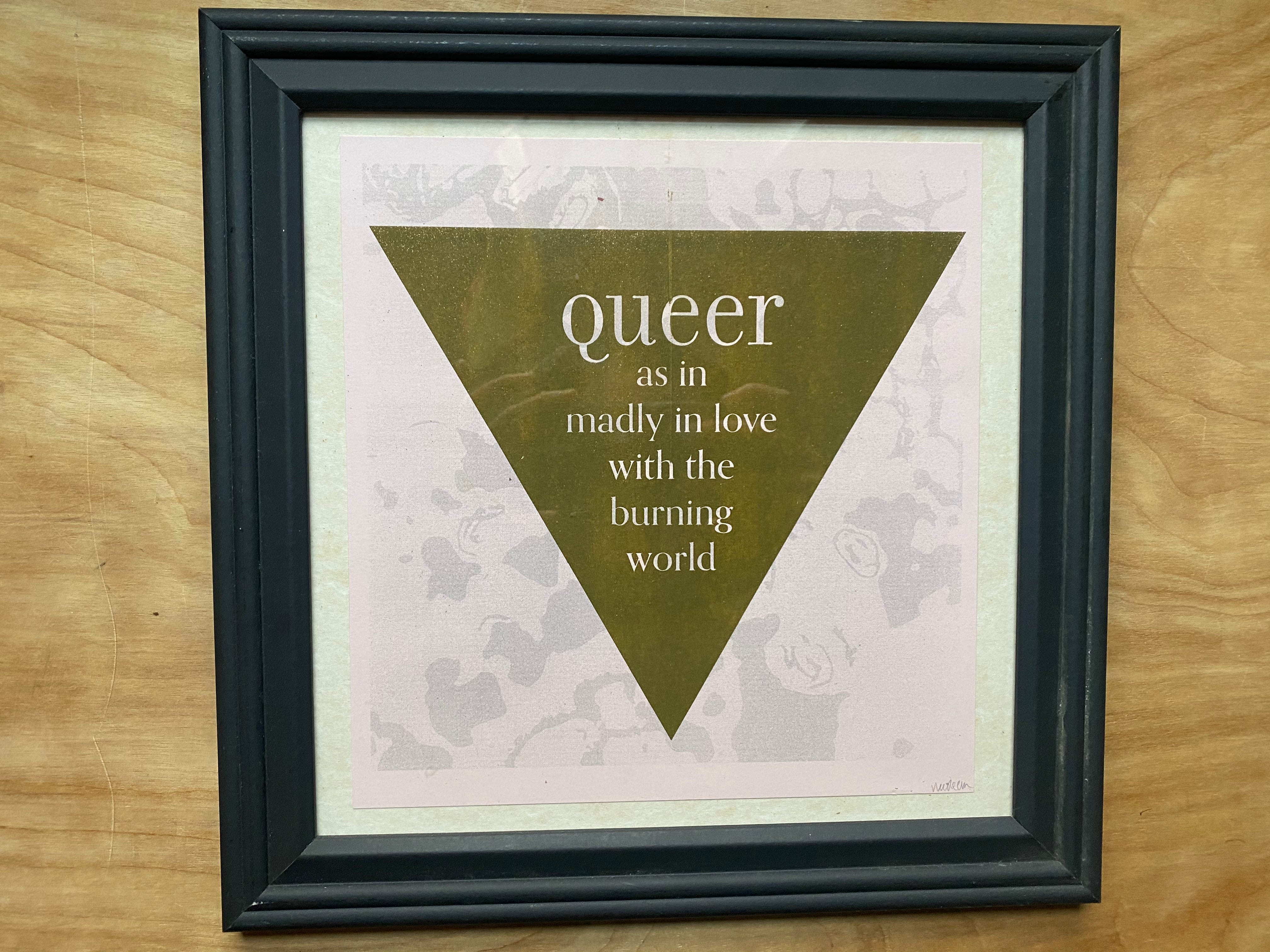 A print in a black square frame. Against a mottled pink and grey background is an upside-down triangle. Inside the triangle are the words: queer as in madly in love with the burning world. “Queer” is bigger than the rest of the text.