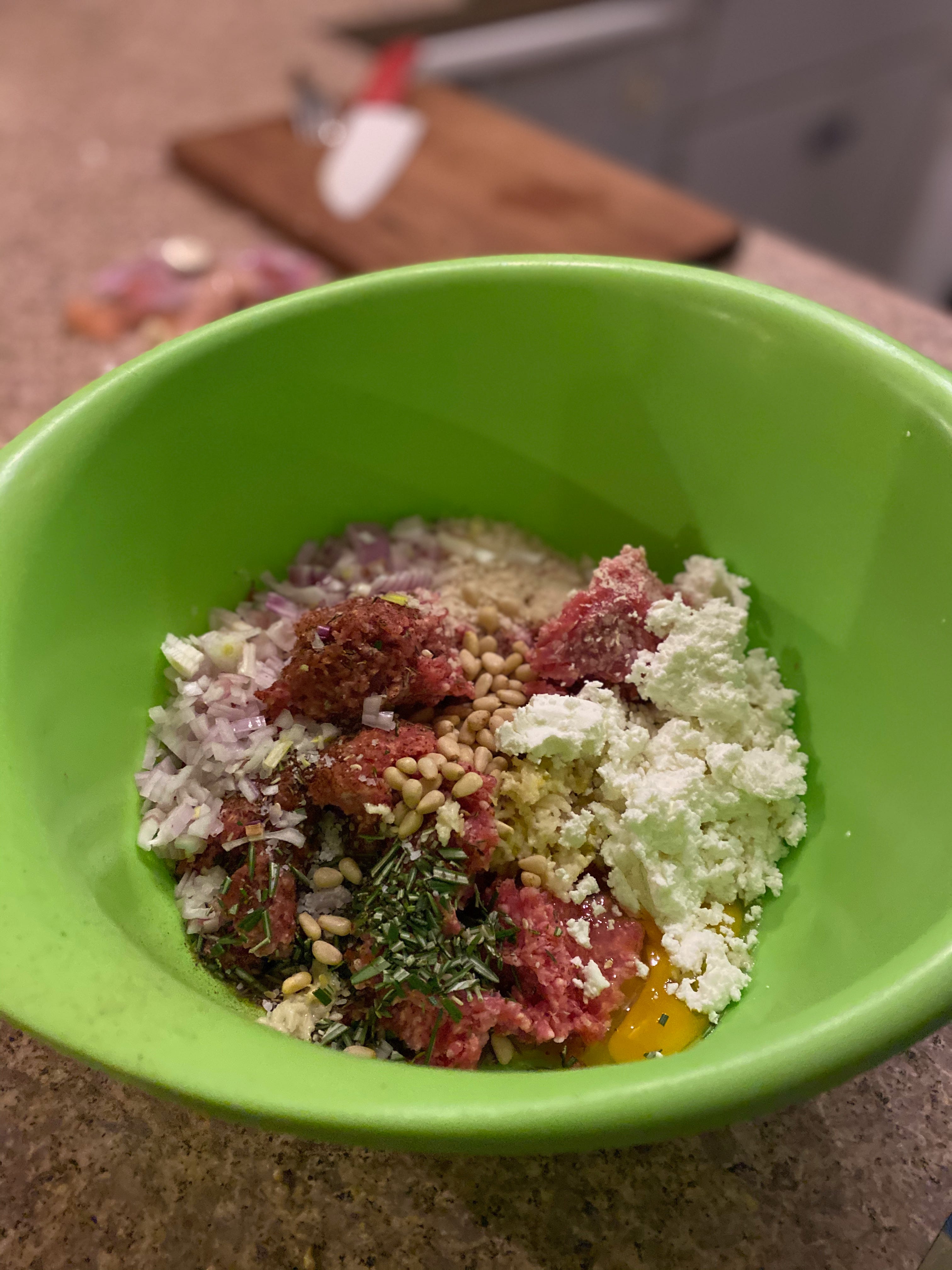A green mixing bowl containing the raw ingredients for meatballs in small piles: diced onion, pine nuts, minced rosemary, an egg, bread crumbs, crumbled feta cheese, and ground lamb.