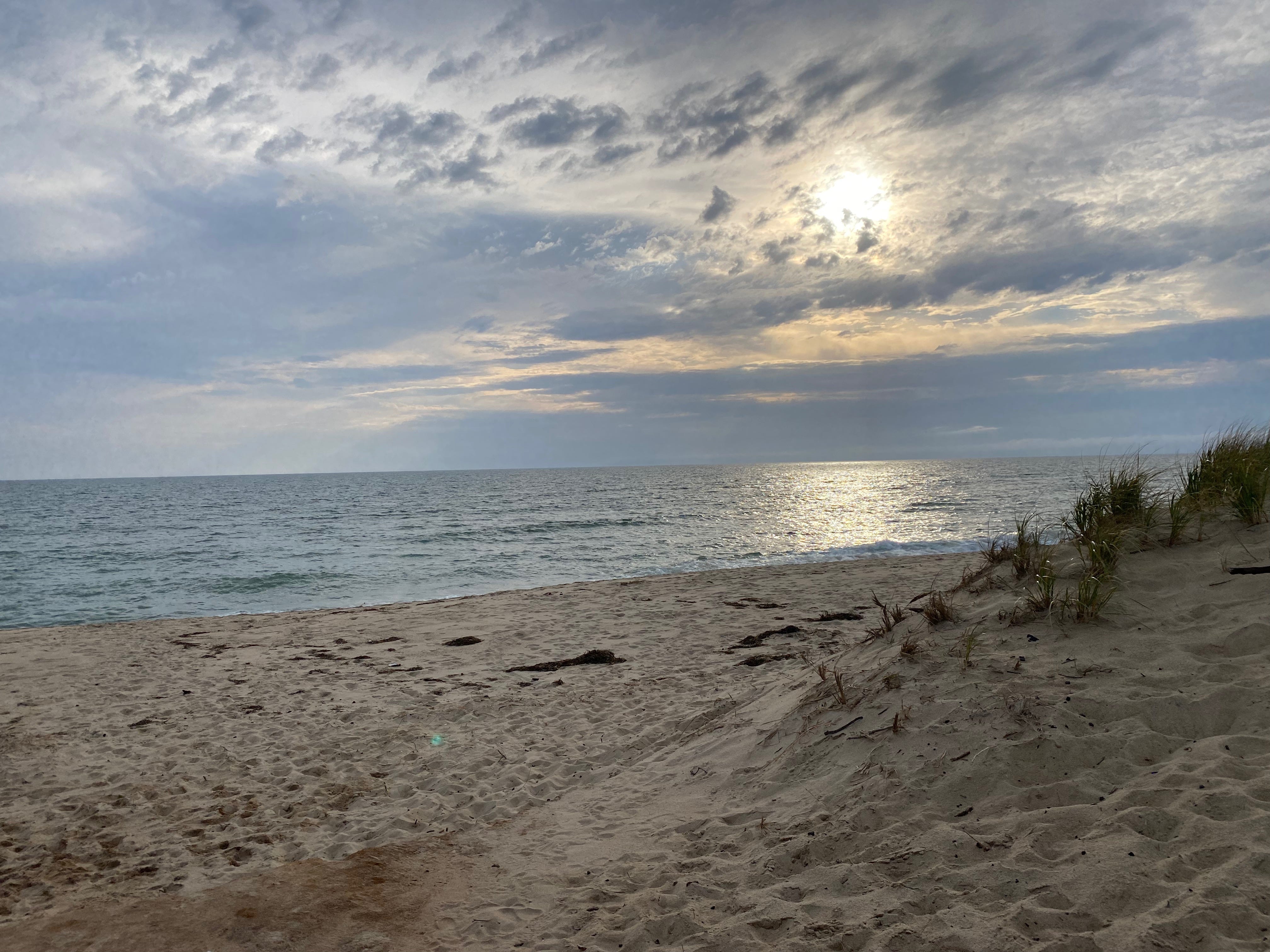 View of a beach in the late afternoon; the sun shines golden on a dark ocean through a sky of blue and grey clouds.
