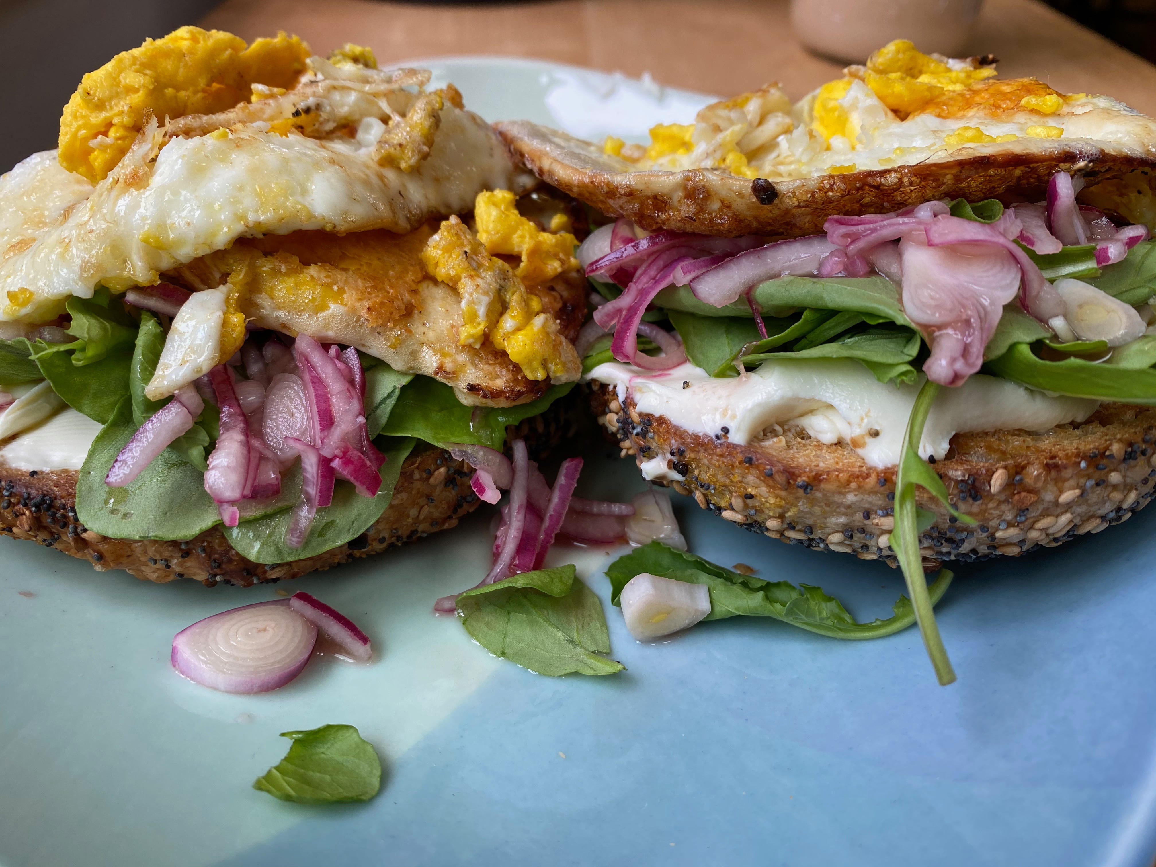 Close-up of two bagel halves on a blue plate, topped with cream cheese, spinach leaves, picked red onions, and fried eggs.