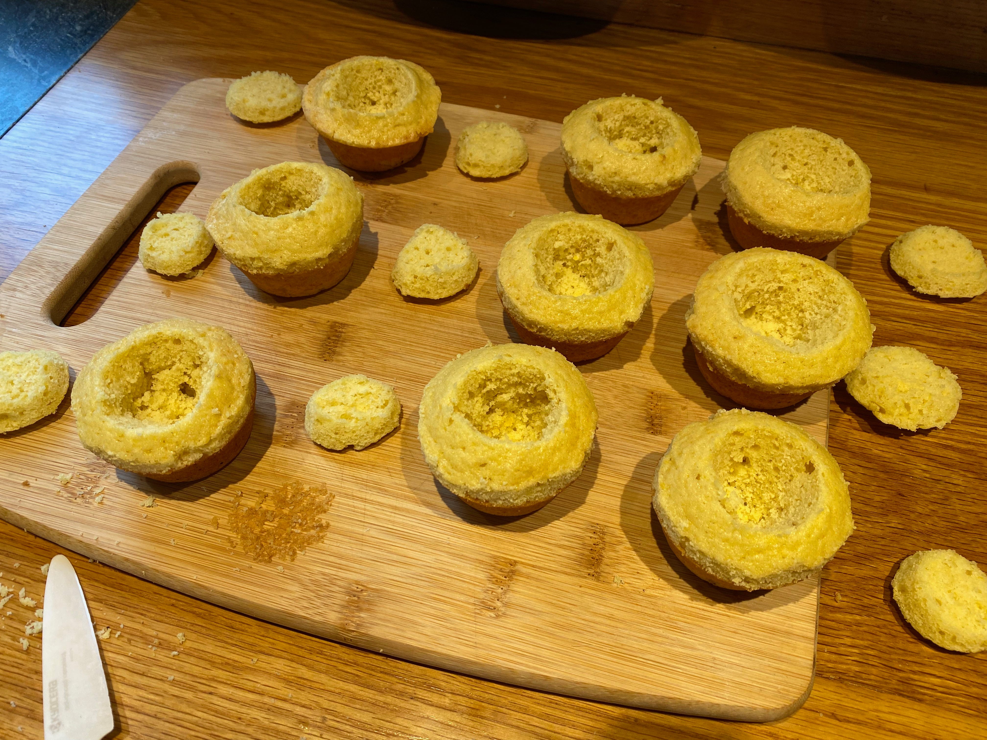 Nine cupcakes arranged on a wooden cutting board. Their centers are hollowed out, and the small pieces that were cut away from them are sitting next to each cupcake, ready to be reassembled.