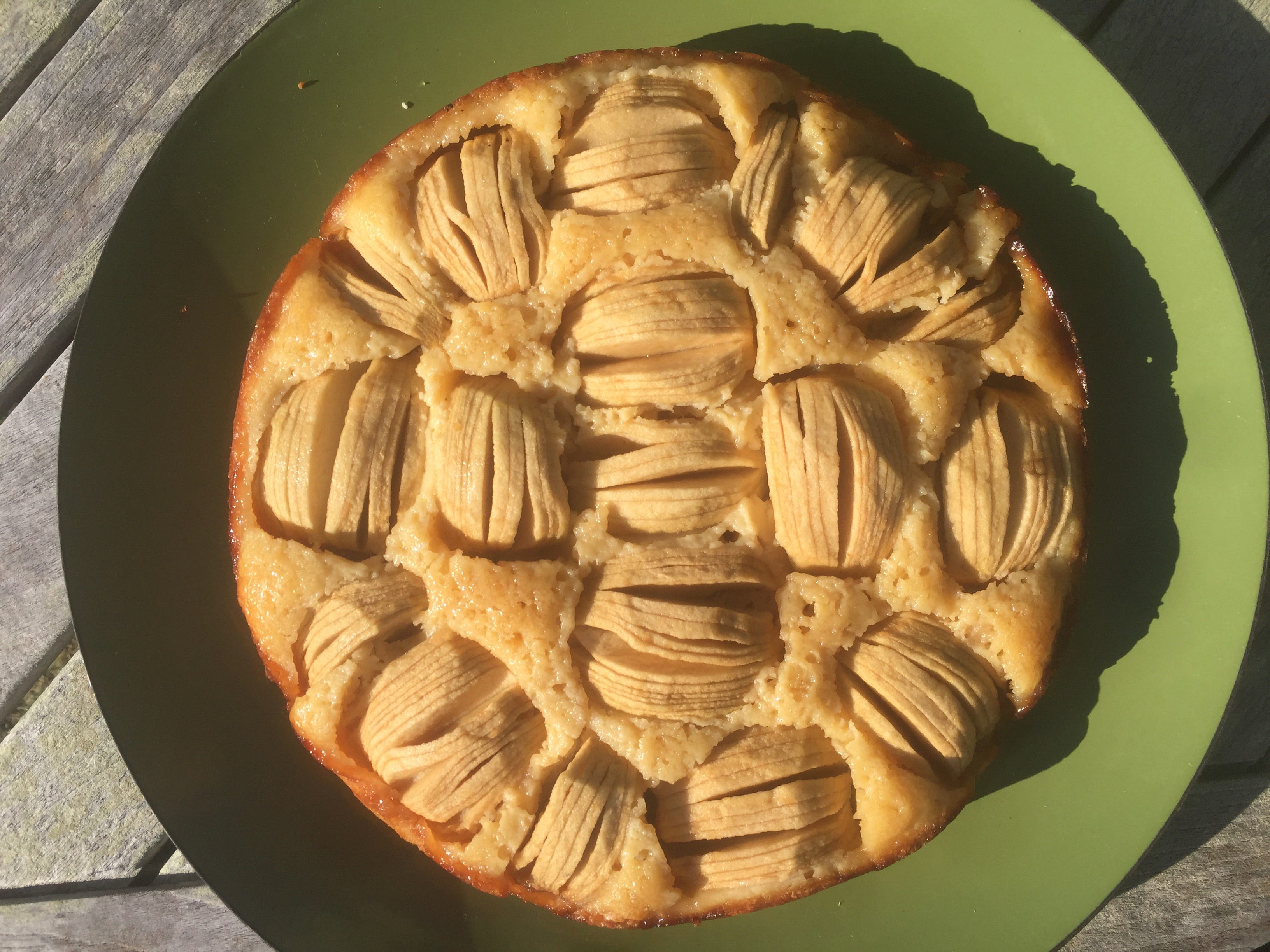 A round cake sits on a green platter. The top of the cake is studded with apple halves, cut into silvers and slighted fanned out into half-domes.