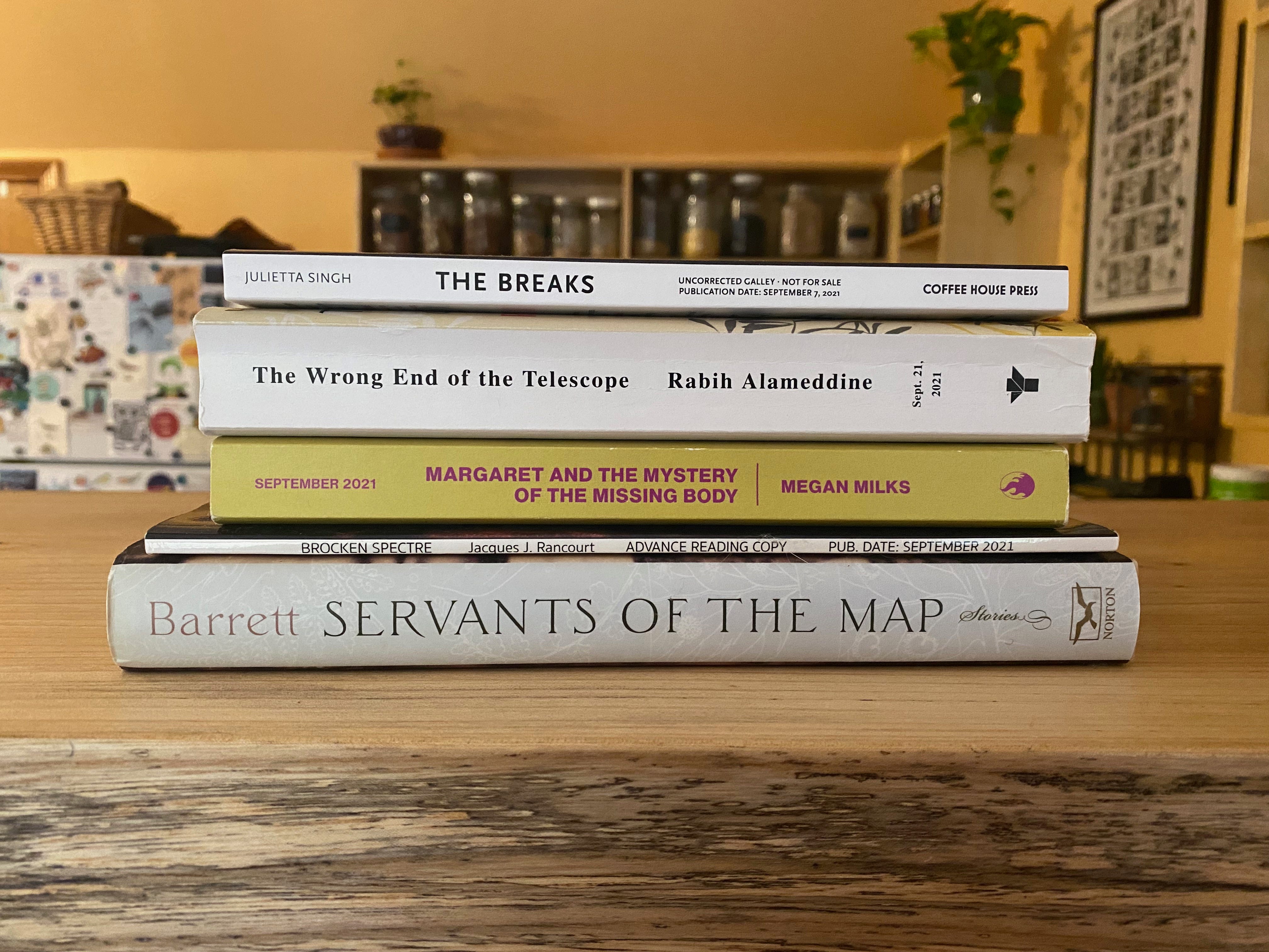 A stack of the following books on a kitchen counter, with a view of pantry shelves and a fridge in the background: The Breaks by Julietta Singh, The Wrong End of the Telescope by Rabih Alameddine, Margaret and the Mystery of the Missing Body by Megan Milks, Broken Spectre by Jacques J. Rancourt, Servants of the Map of Andrea Barrett.