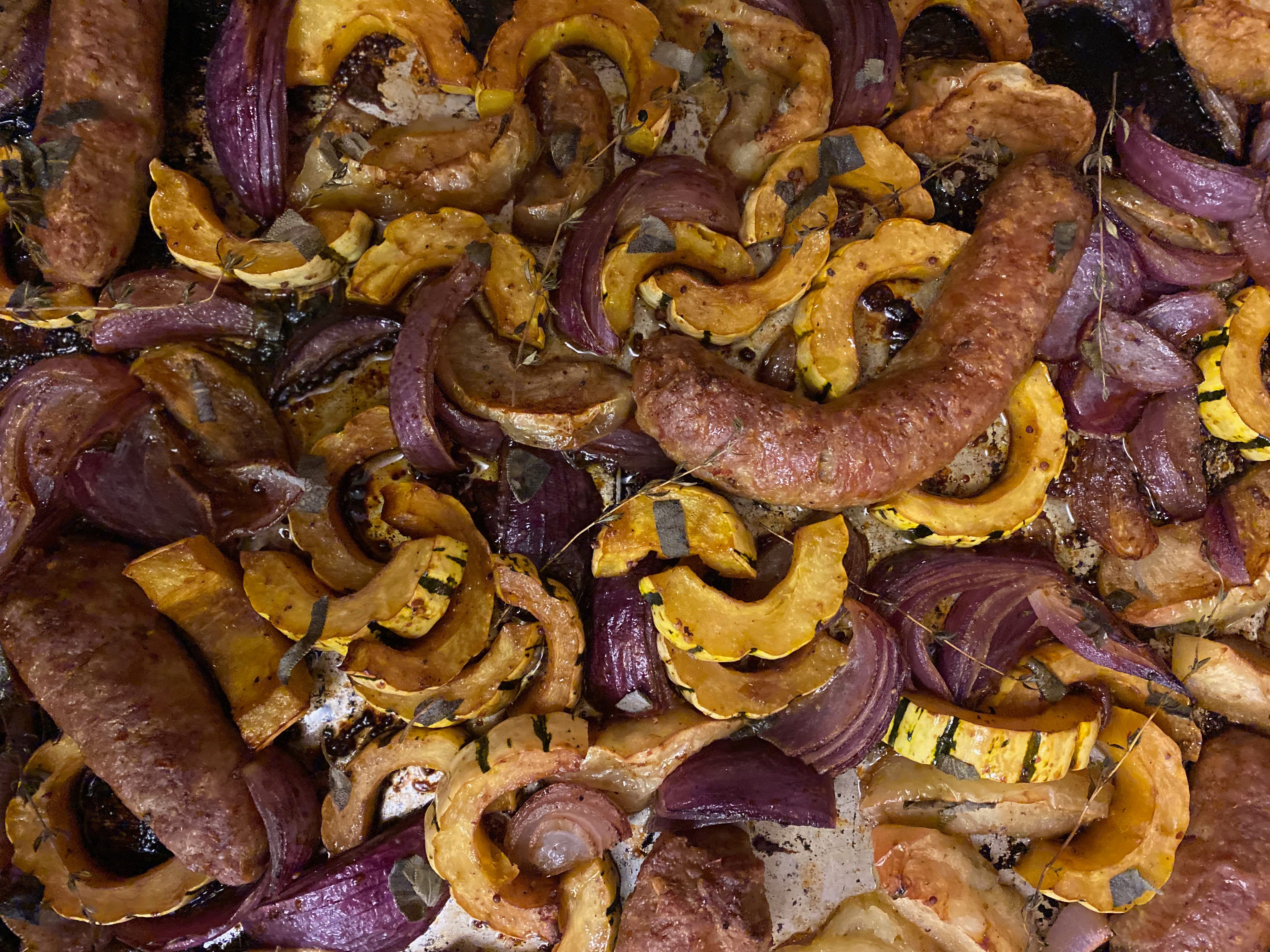 Close up of a pan of roasted half-moons of delicata squash, wedges of red onion, and soft apple quarters nestled among juicy sausage links.