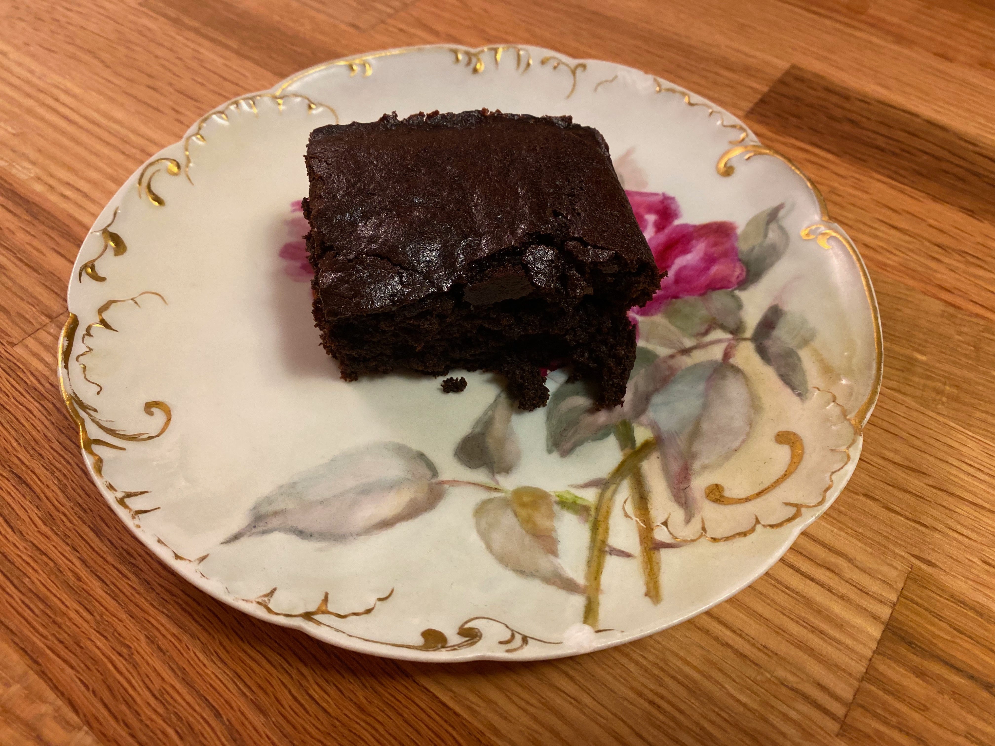 A square of very dark gingerbread sits on a painted ceramic plate on a wooden counter.
