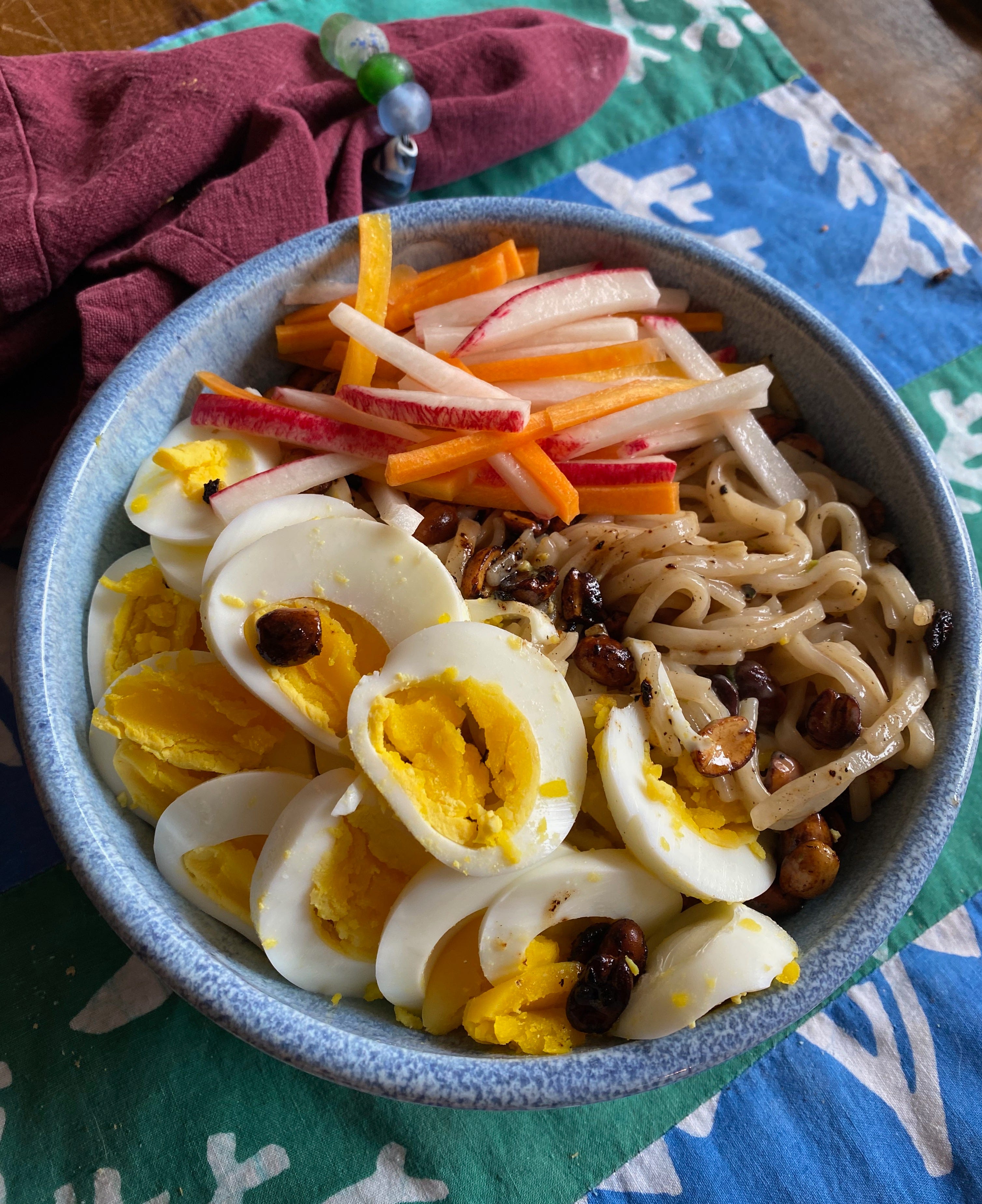 A ceramic bowl full of rice noodles, sliced hard-boiled egg, thin matchsticks of picked carrot and daikon, and spicy fried peanuts.