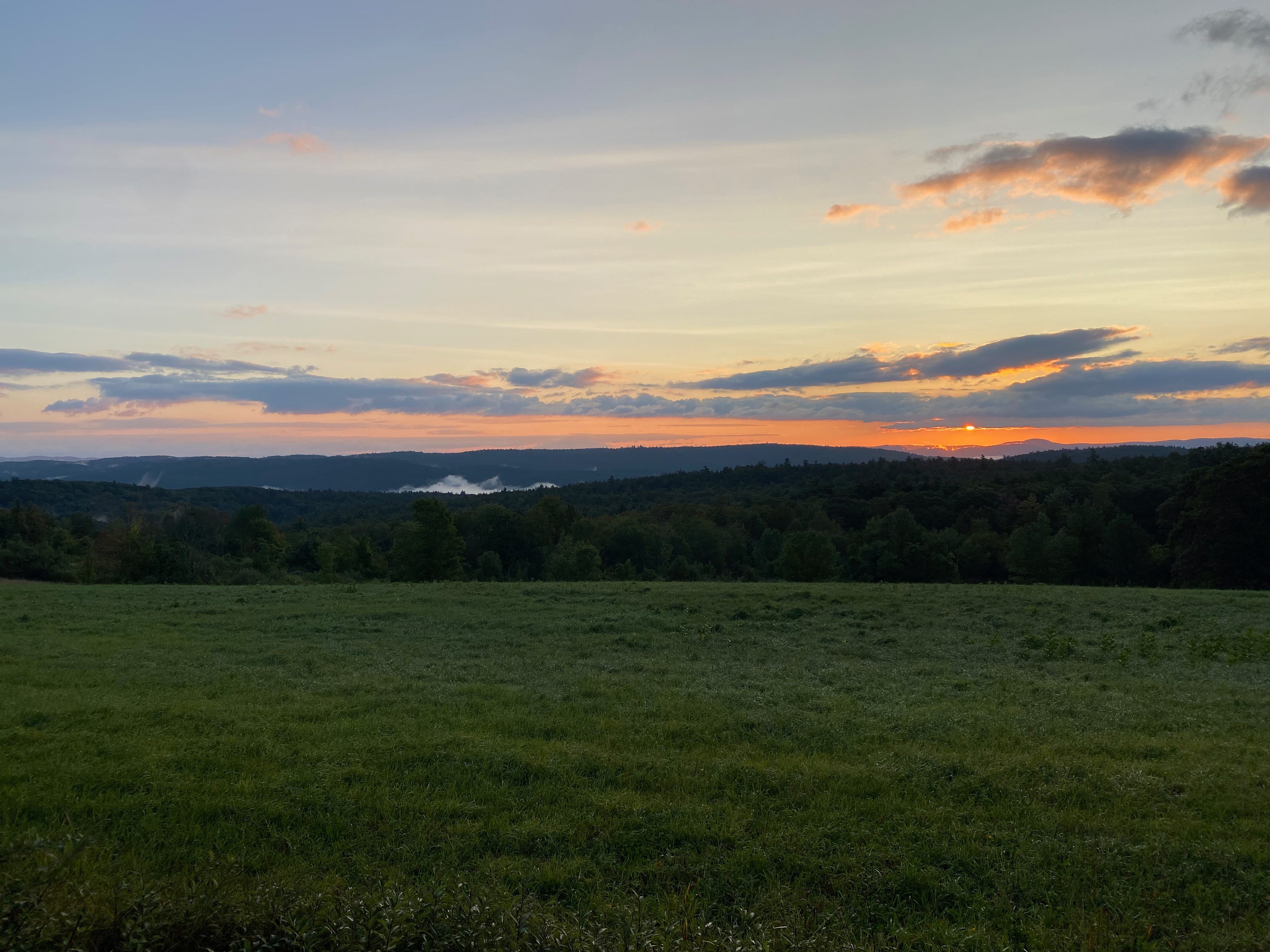 Sunrise over a grassy ridgetop pasture. Wisps of clouds sit in the mountains and the sun is a tiny golden orb among purple and orange clouds.