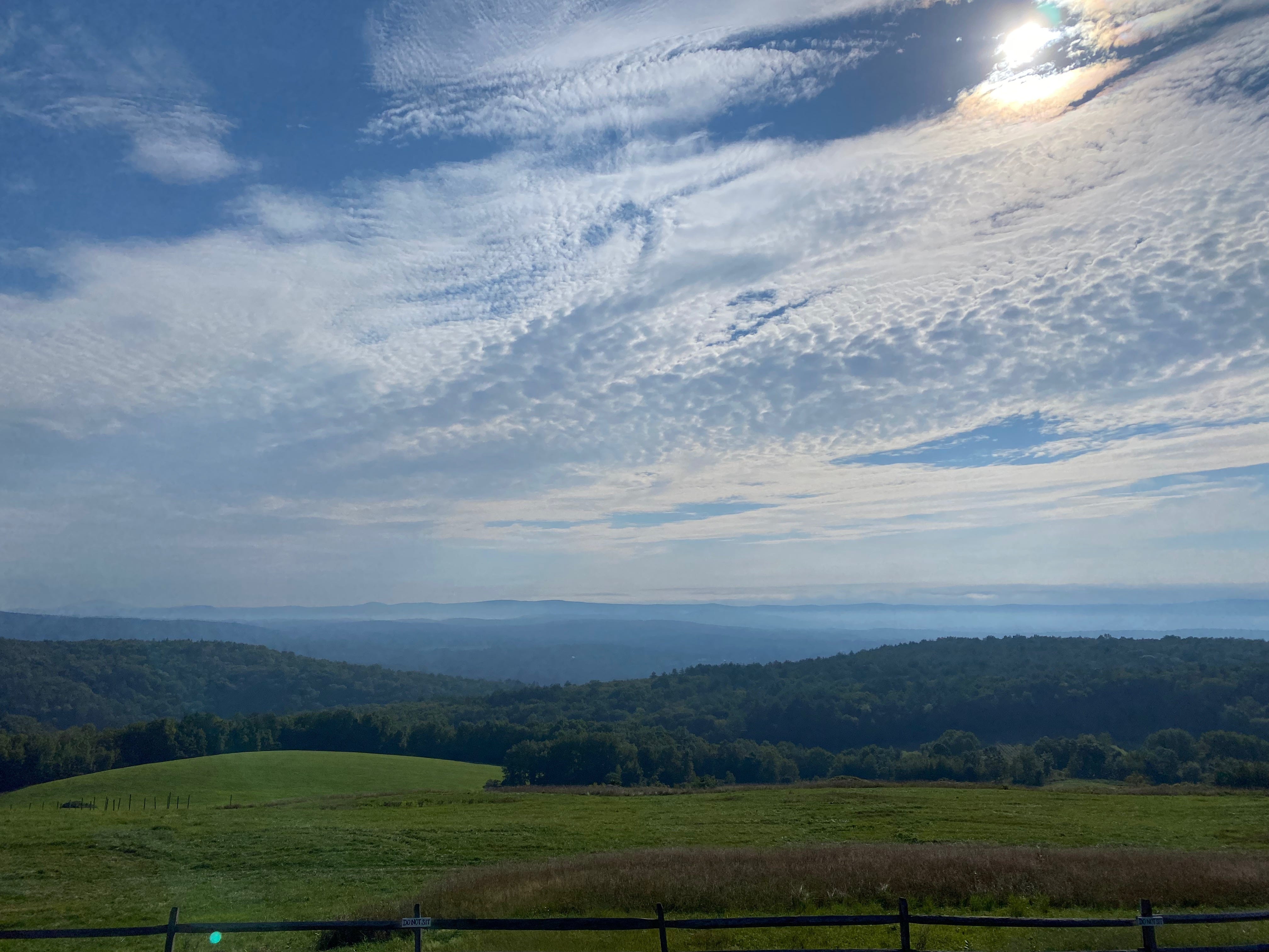 View of a sloping green field, looking out to hazy hills and mountains, under a wide sky of white and silver clouds.