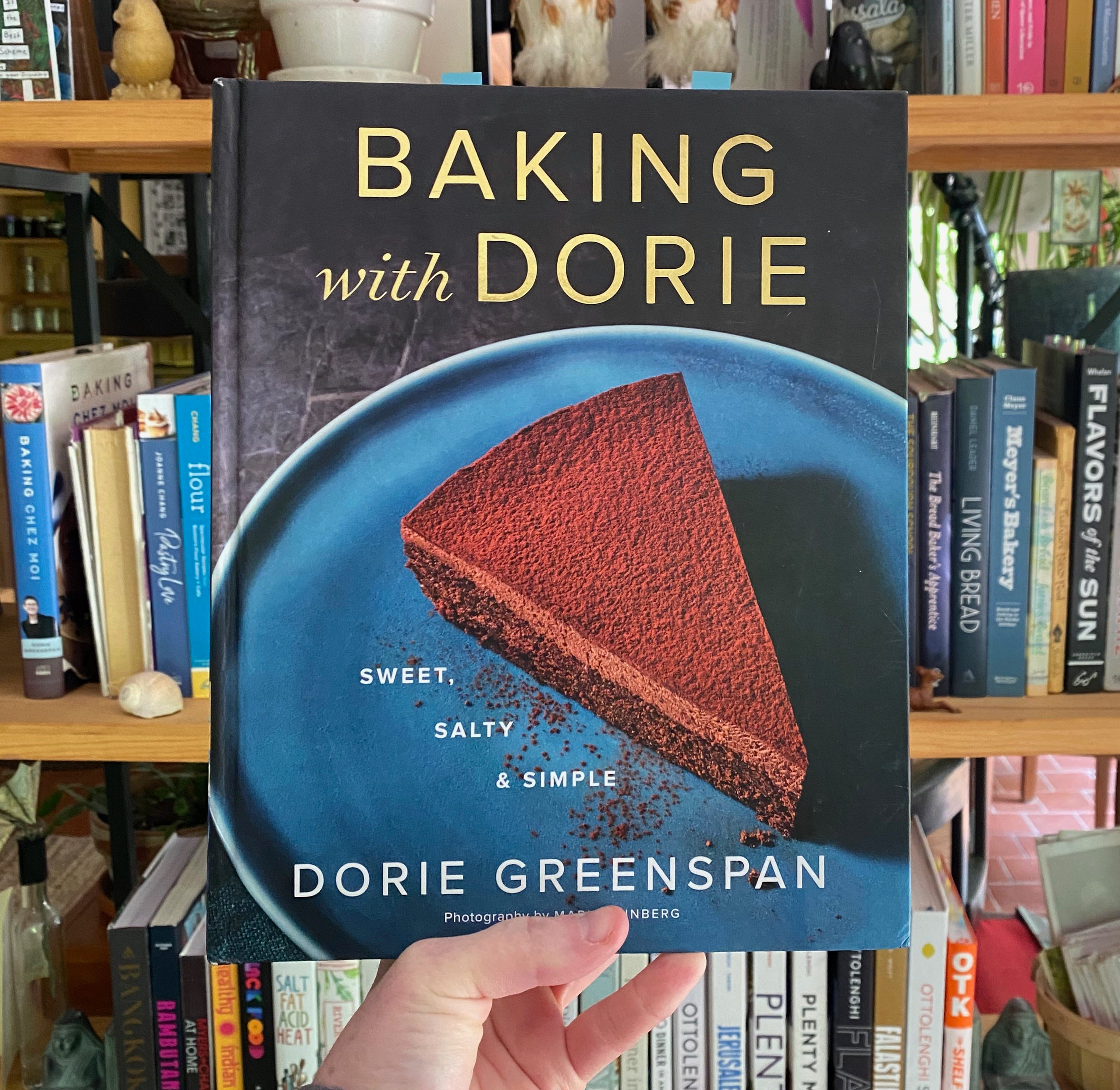 My hand holding this cookbook in front of a shelf full of colorful cookbooks.