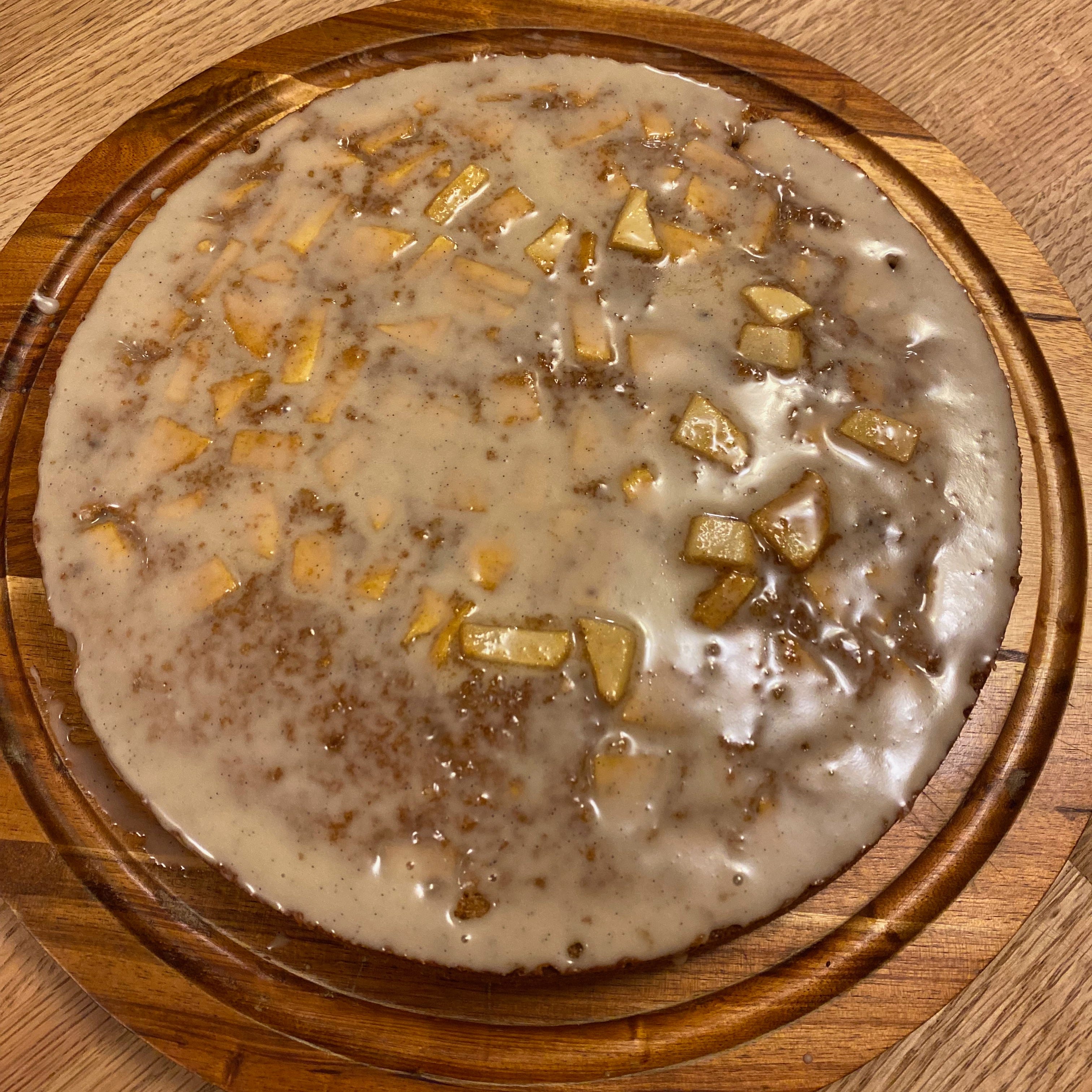 A round cake covered in a shiny white glaze and studded with chunks of apple, sits on a wooden cake plate. 
