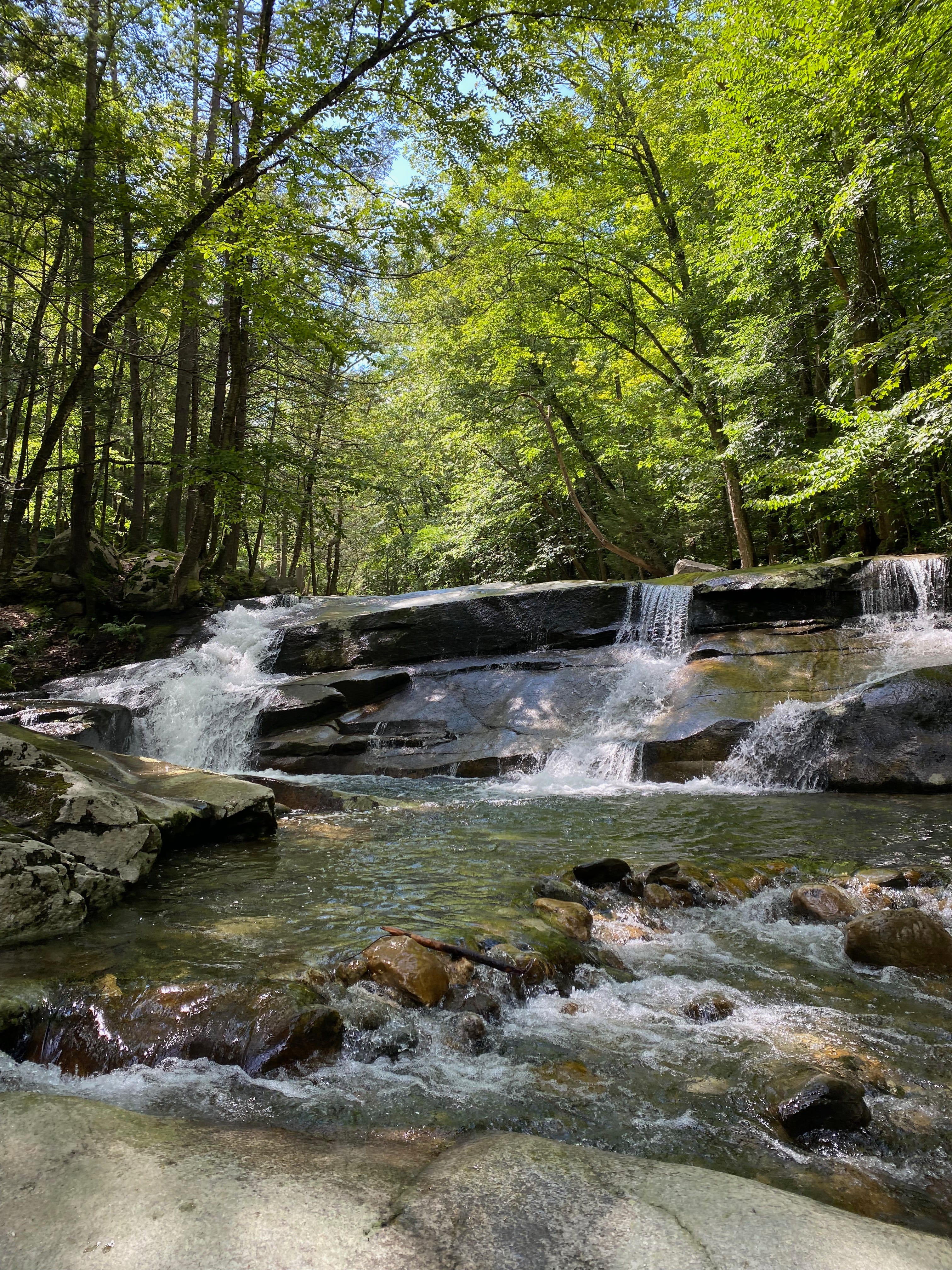 A series of small, frothy, glittering waterfalls cascading over smooth rocks into a deep pool on a sunlight river, surrounded by green trees and patches of blue sky.