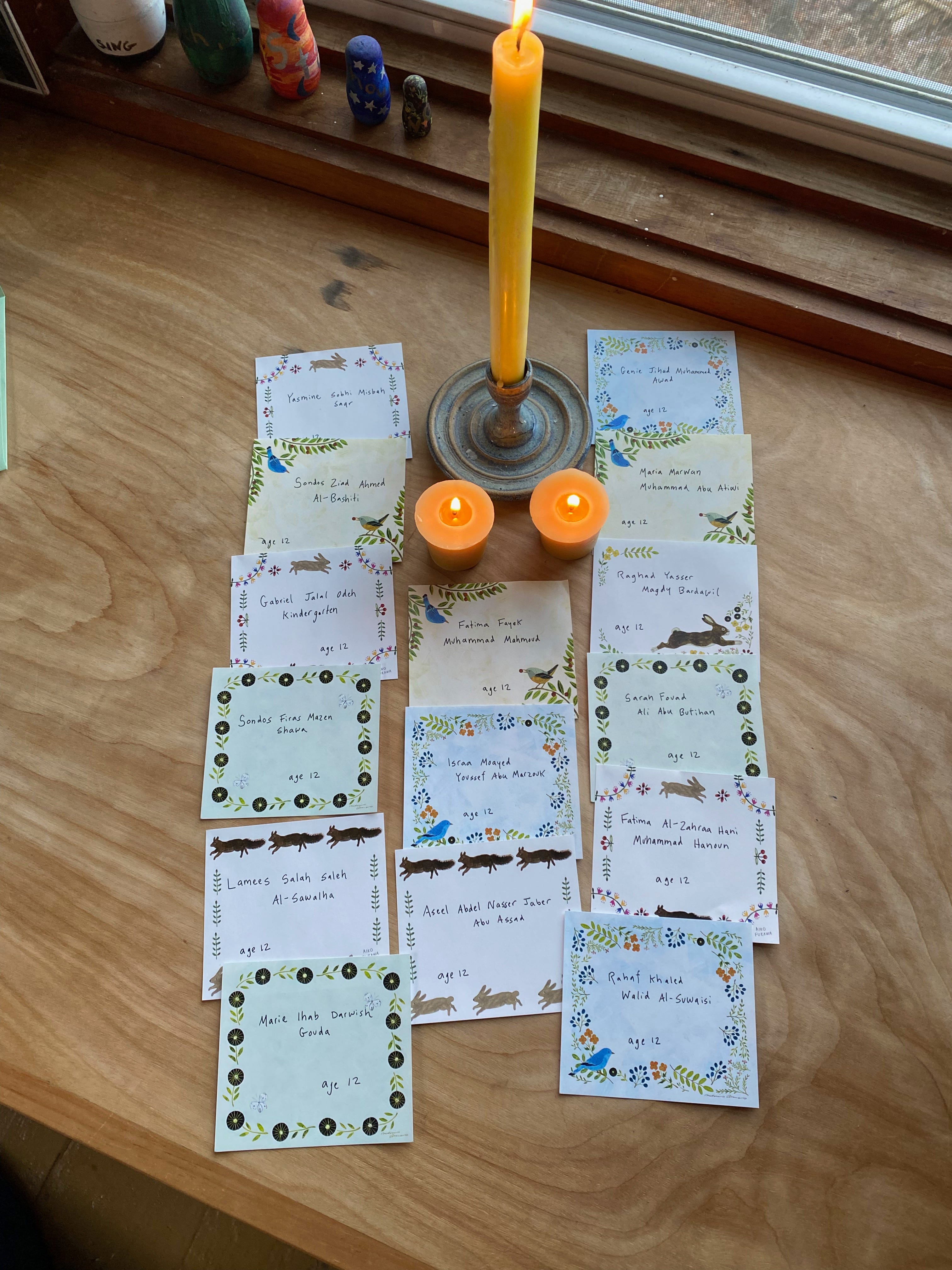 15 small slips of stationery, each with a handwritten name, surround one tall candle and two smaller ones. 