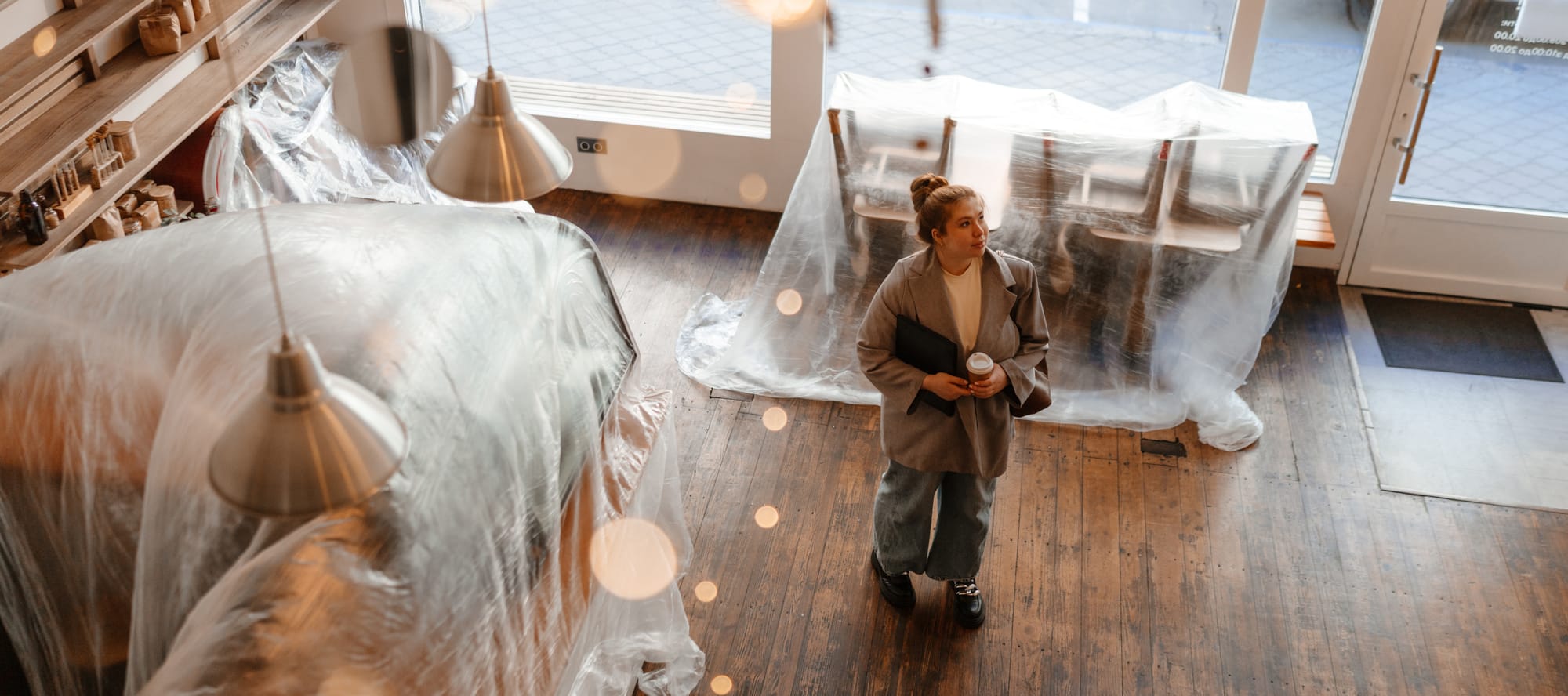 A White young female cafe owner is standing in her cafe, holding a coffee cup and a laptop, surrounded by plastic covers on furniture