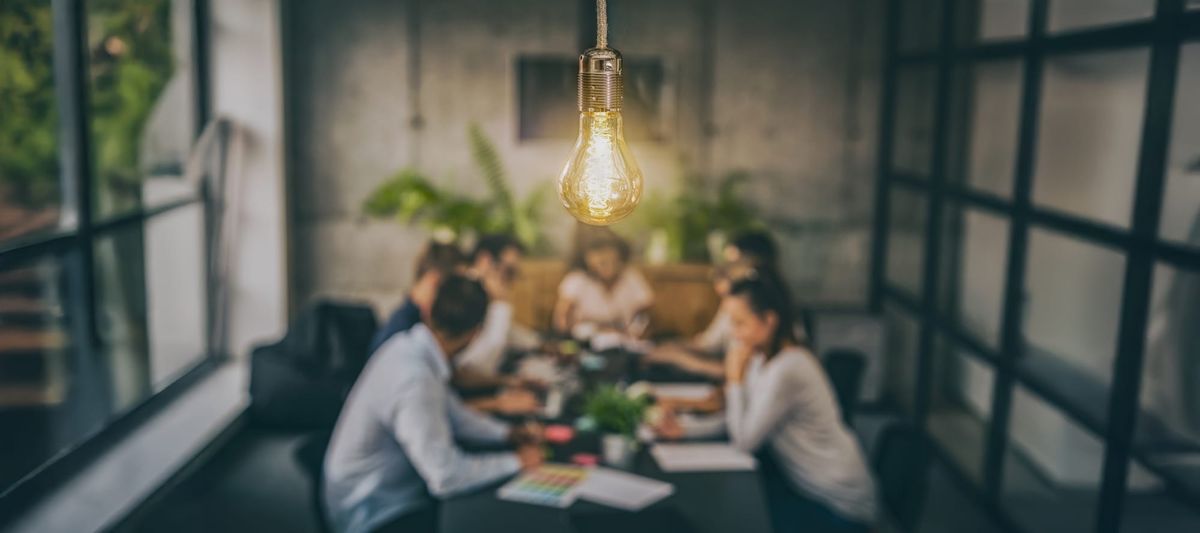 A group of people working together at a table in an office. A lit lightbulb shows in the foreground.