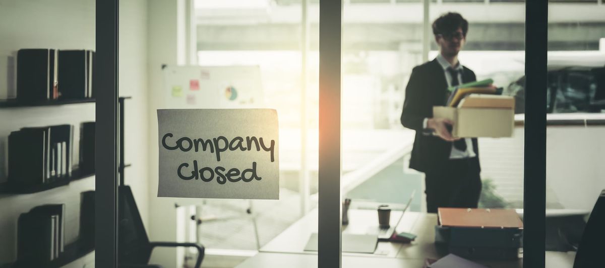 Company closed on windows of an empty office with business man packing belongings walking out of business