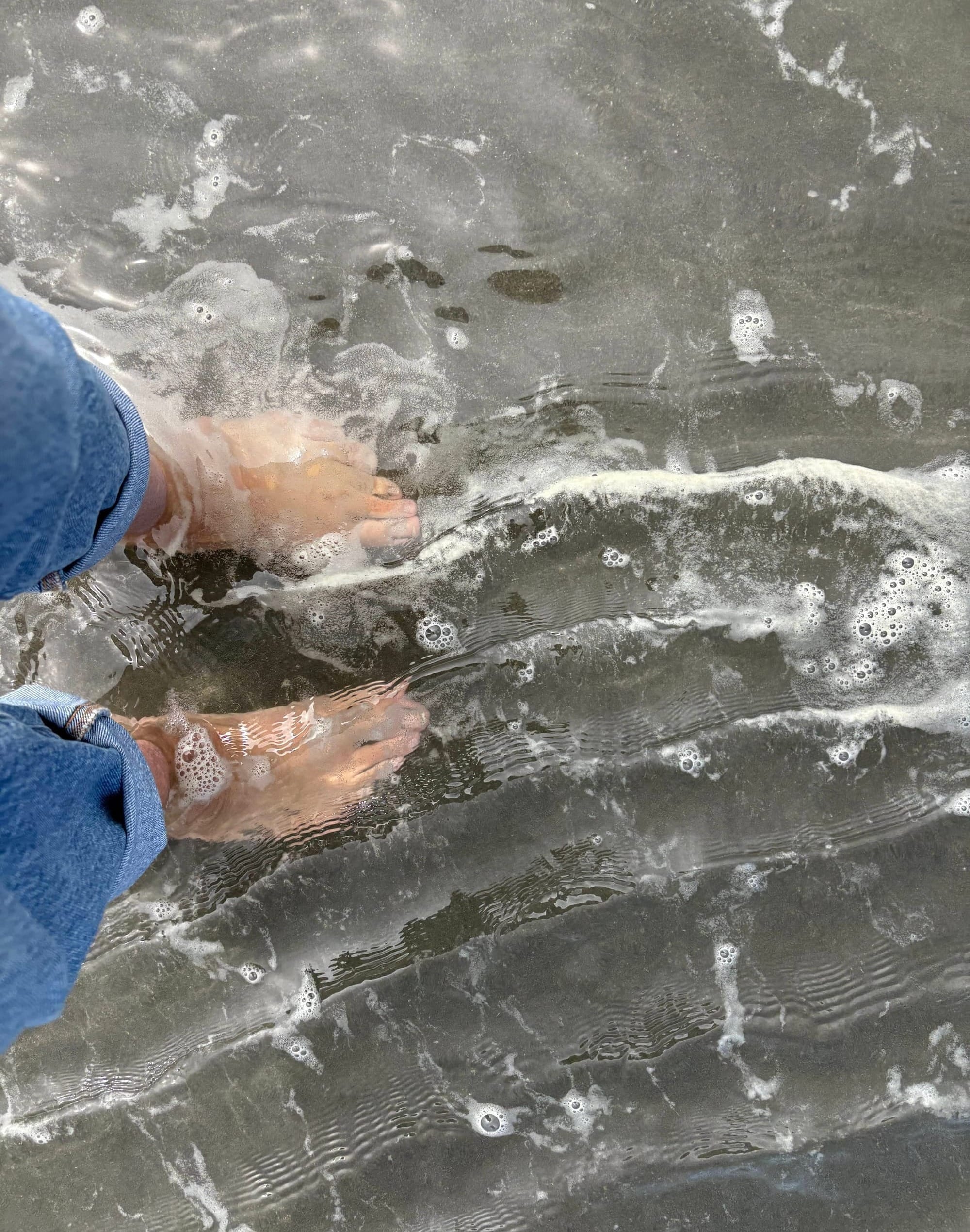 Photo of are feet, rolled up jeans, in the shallow ebb of Pacific surf.