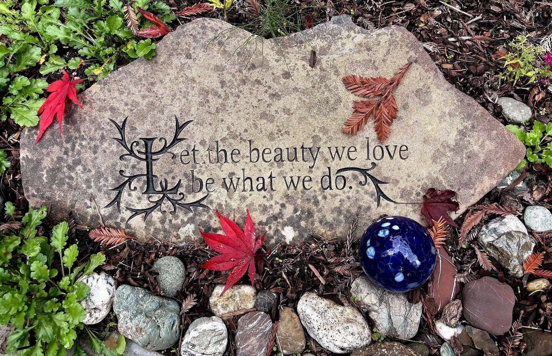 Photo of a carved stone that says, Let the beauty we love be what we do, with stones, and two small Japanese red maple leaves.