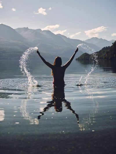 Photo of woman waist-deep in river flinging water joyfulling into air, reflected in water.