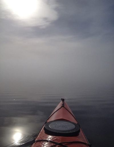 Photo of front tip of orange kayak on calm lagoon with the sun and bright blur in the clouds and reflected on the water.